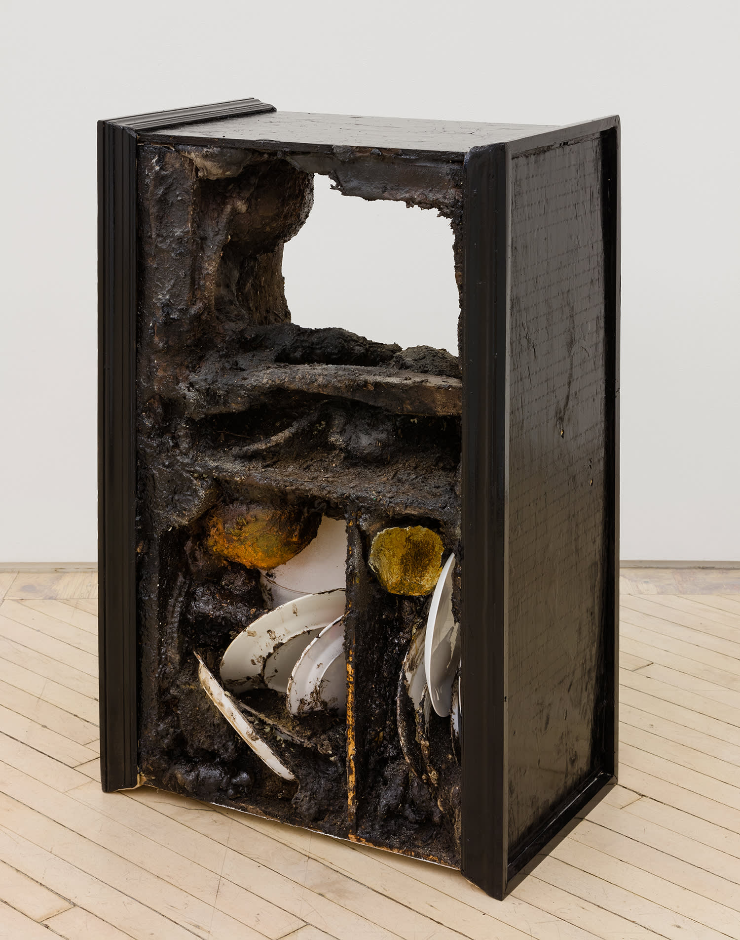Image of a sculpture by Brandon Ndife. Freestanding in a gallery is what appears to be a piece of furniture, like a shelf or TV console, painted black, with dirt and debris spilling from open cavities. There appears to be dirt covered plates in stacked on the bottom shelf.