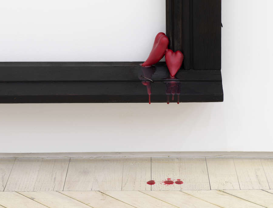 Detail of a wooden frame sculpture, the bottom right angle showing two melting red wax hearts. Some of the wax is dripping down to the white wooden floor.