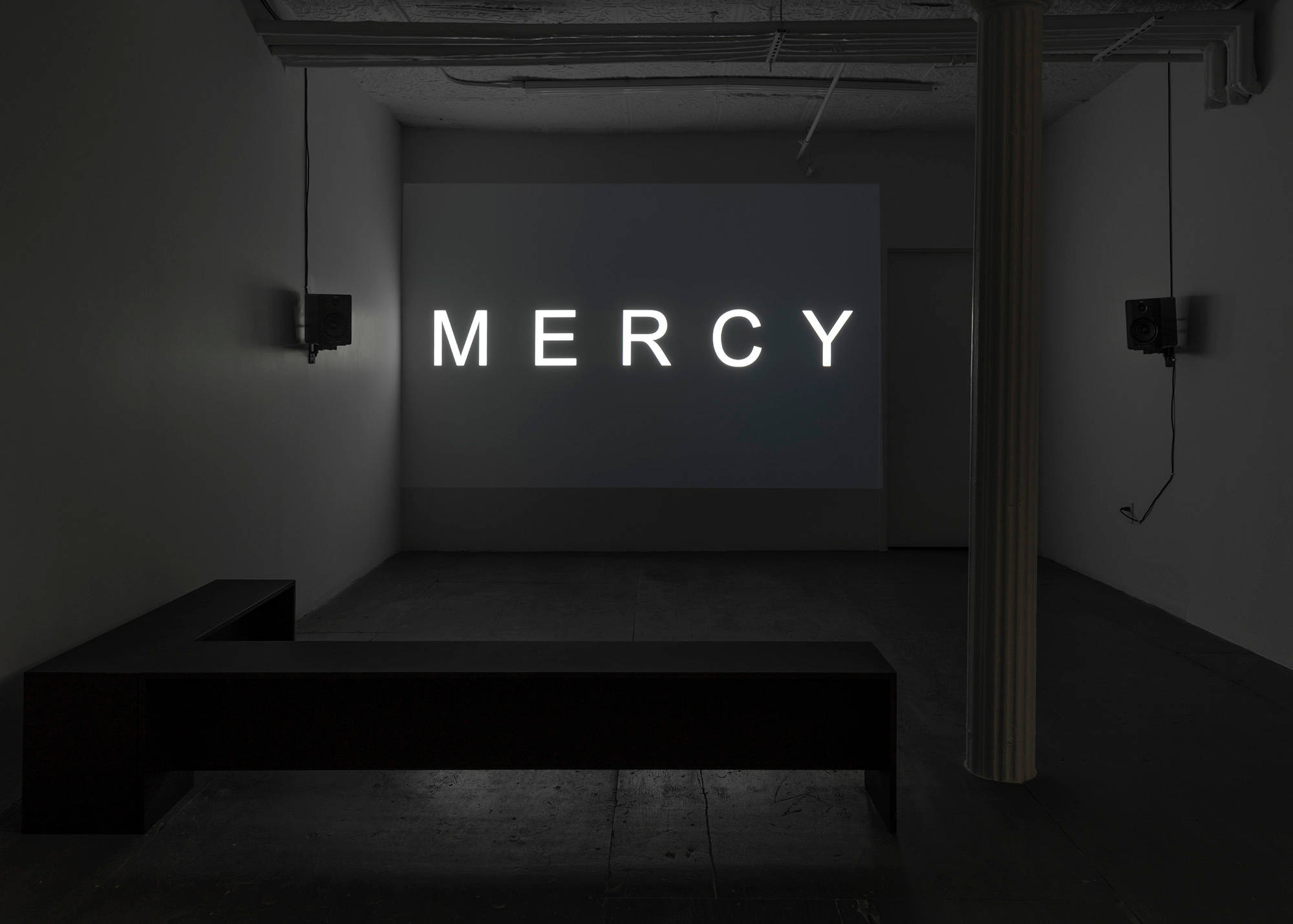 In a dark room, a projector displays the title of the film "MERCY". There are two black benches in the shape of an "L" on the floor of the room. Two speakers hang on opposite walls to the left and right of the screen with cords running up to the ceiling.