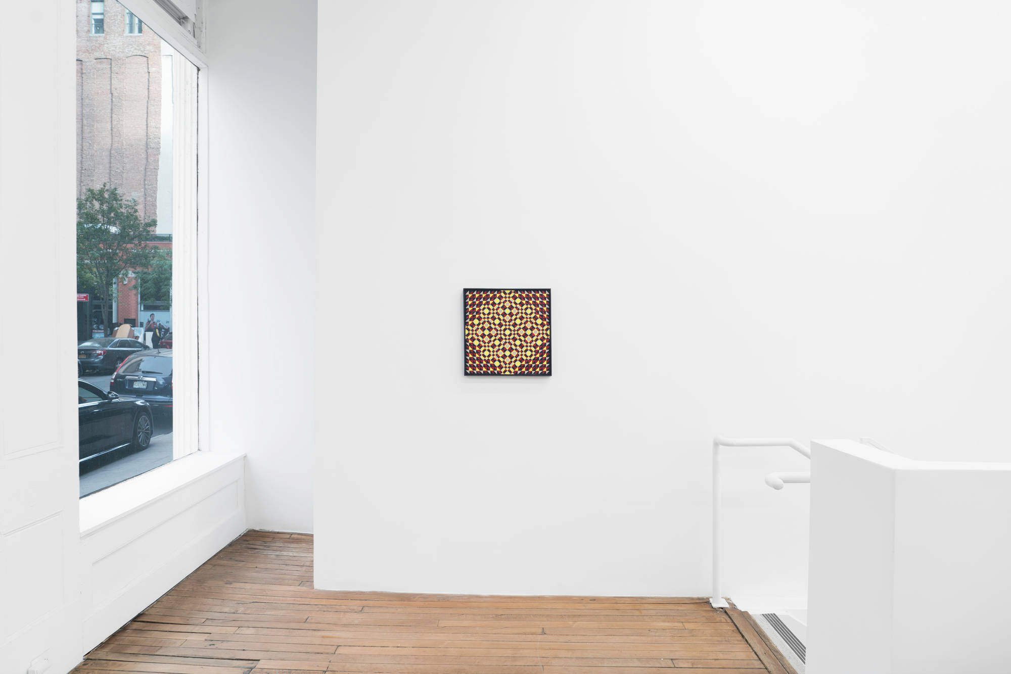 In the north east corner of the gallery, an abstract, geometric painting by Matt Hoyt hangs on the wall. A stairwell descends to the right, out of frame. A view of the street is visible from the window to the left of the painting.