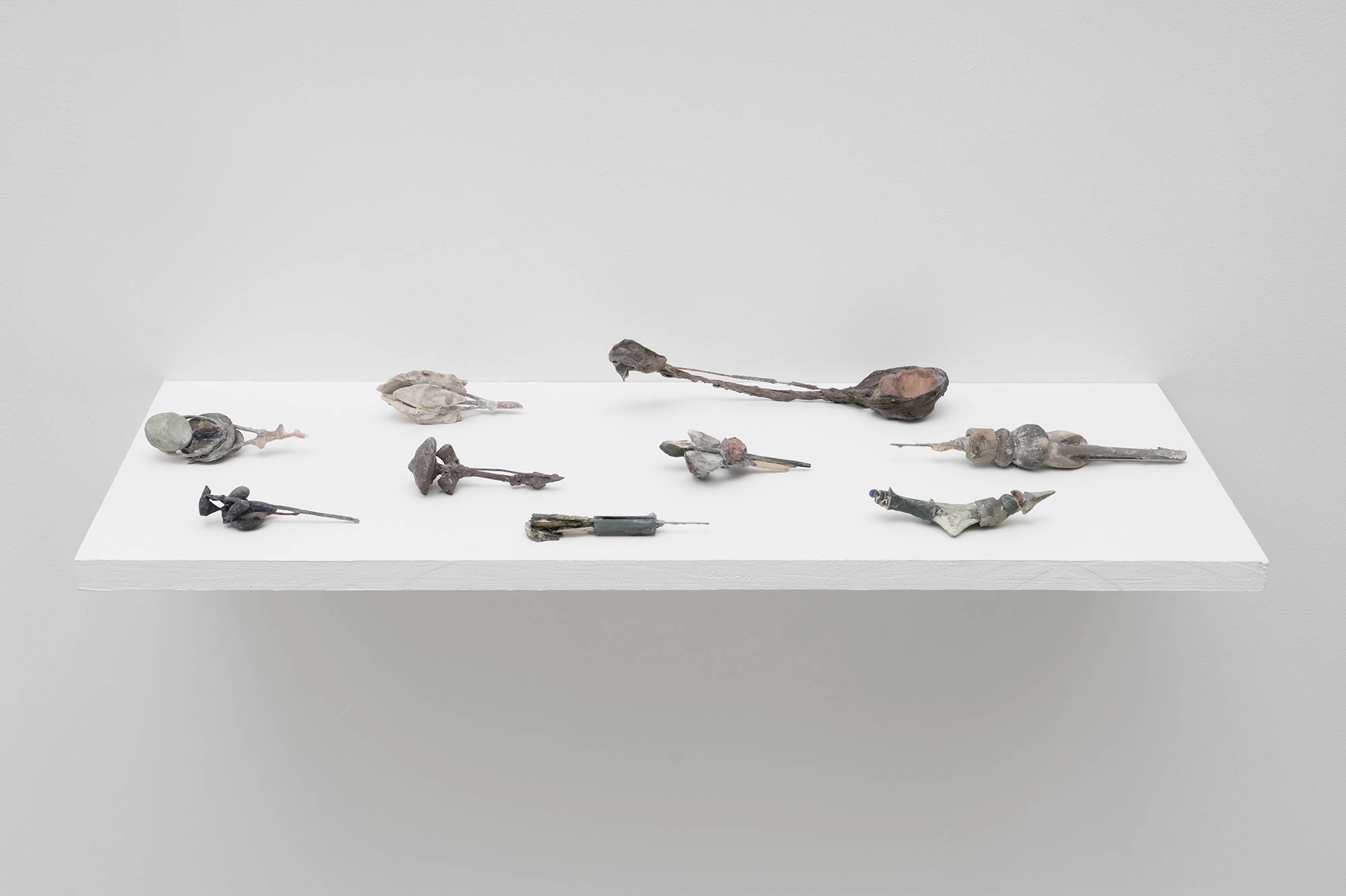 A group of small, intricate, handmade sculptures by Matt Hoyt sit on a white ply wood shelf. The colors are earthy, cool, and muted greens, reds, and greys.
