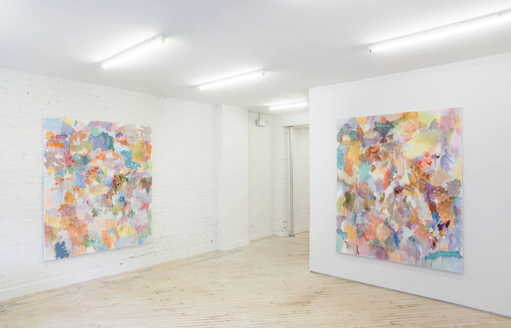 In a gallery space, two large abstract paintings are hung on separate walls. There is a hallway between them. The paintings contain numerous swatches of various color painted on top of one another. The paint is smeared in dripping.