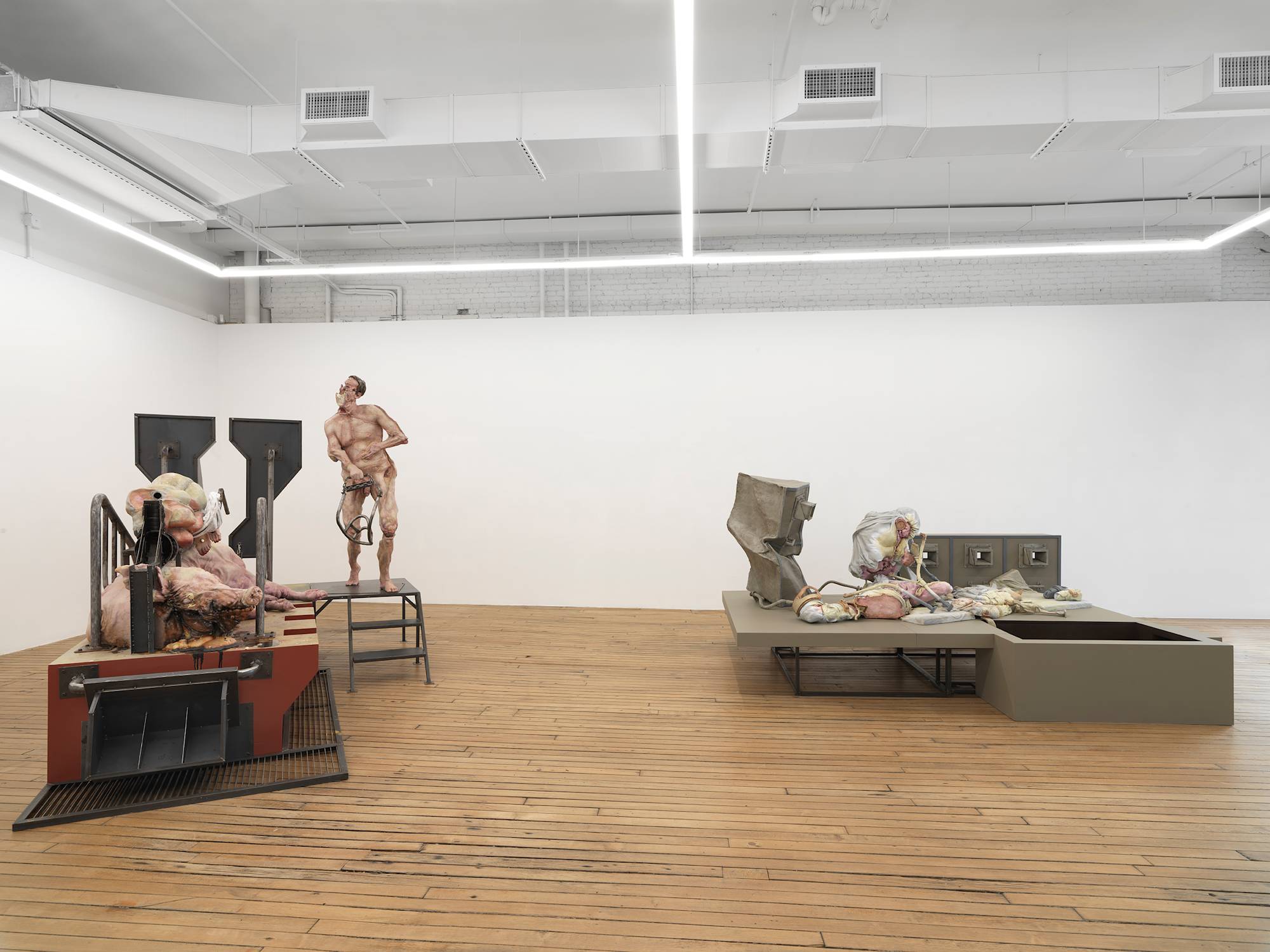 In a large gallery space, two monumental sculptures are spaced apart. Both sculptures contain a combination of abstracted body parts and steel structures raised off the ground.