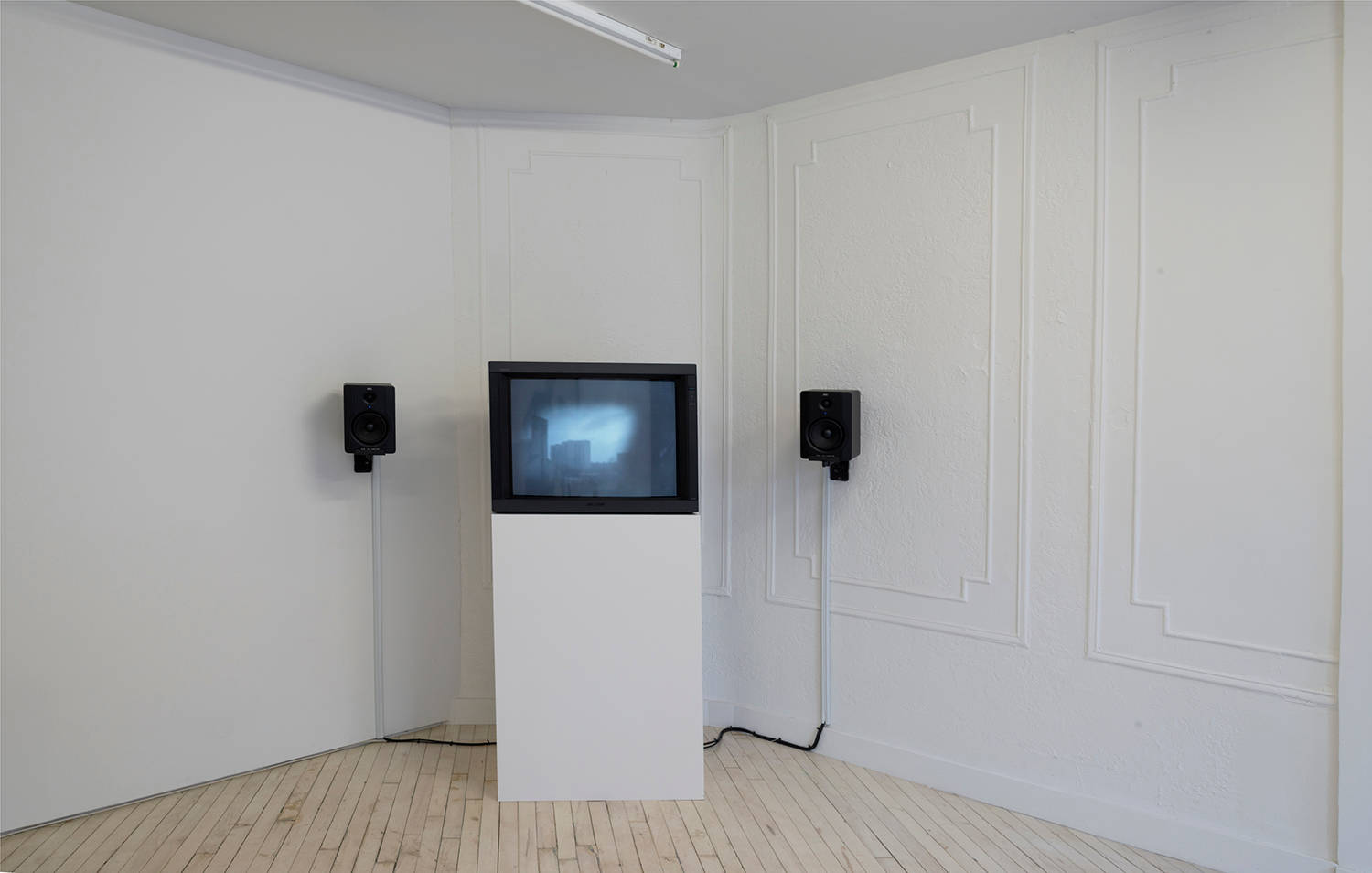 Installation view of a tv monitor with 2 black speakers on either side, on a white plinth - the monitor shows a blurred black and white image of a wojciech bakowski video.