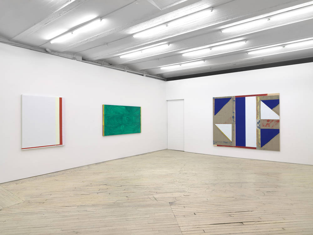 In a large gallery space, three abstract paintings in a line. On the left is a white monochrome painting with a red band of color along the right and bottom edges. In the center a horizontal green painting with a brushy surface. To the right a larger abstract painting with shapes painted in blue, white, red. 