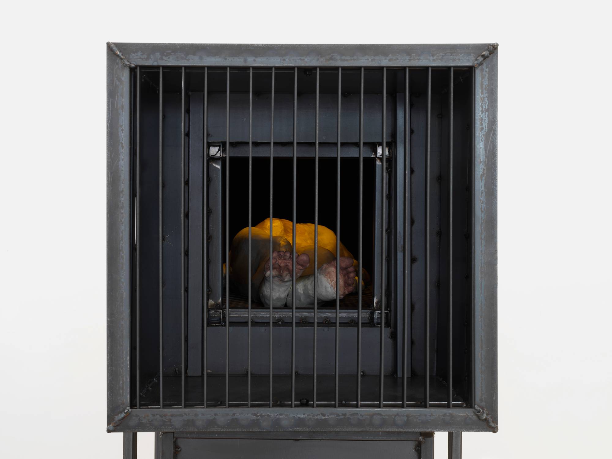 A large steel sculpture with gated bars at the front. Inside there is a form resembling a human laying down underneath a yellow source of light.