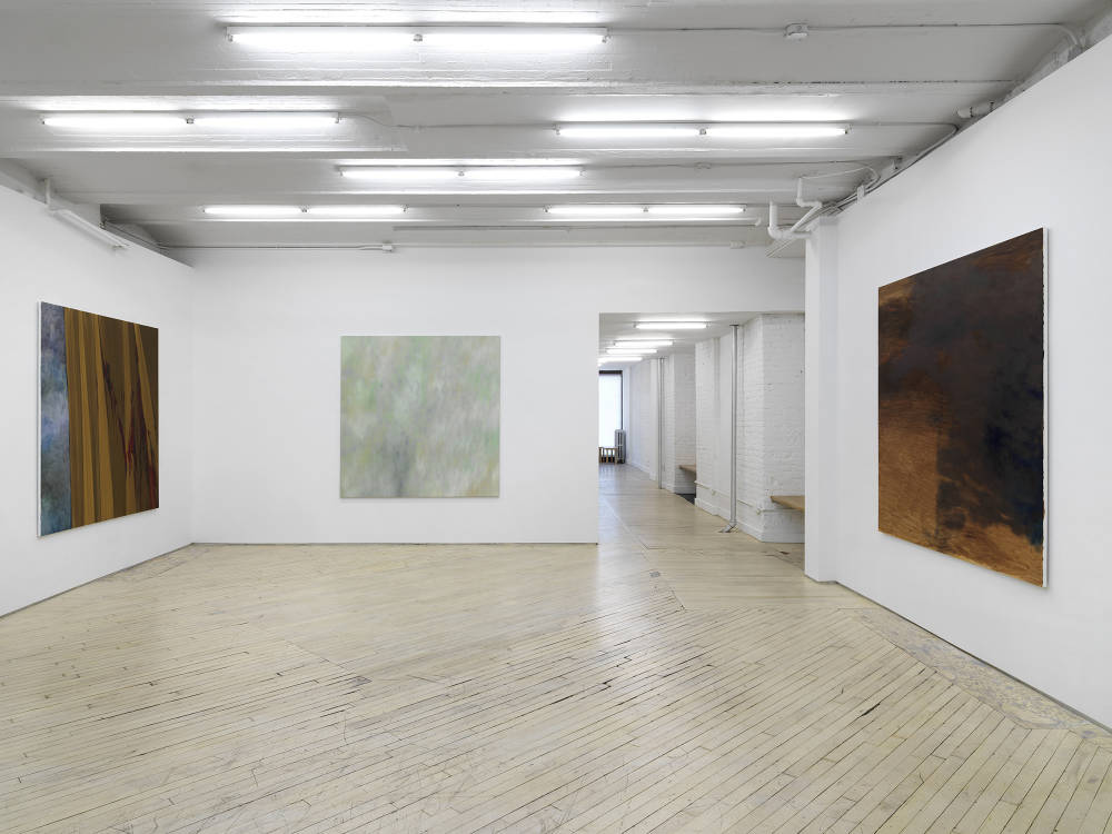 Three large, square abstract paintings installed on separate walls in a large gallery space with the long hallway in the back.