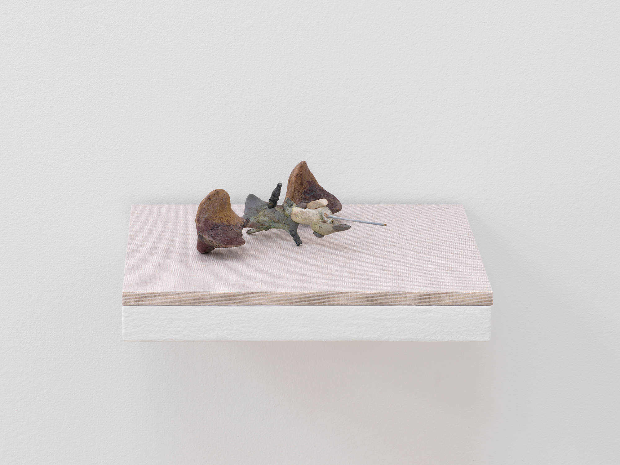 A small abstract sculpture resembling a fossil, bone formation, or combination of organic materials. The object is primarily gray and tan and resting on a shelf with a tan base attached to a white wall.