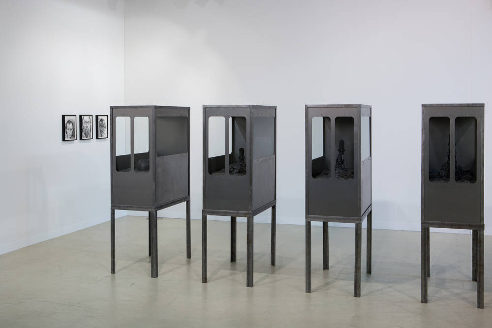 View of 4 standing metal sculptures and at back left 3 small black and white drawings framed in black frames. The 4 structures are identical, rectangular structures with windows made of dark grey steel.