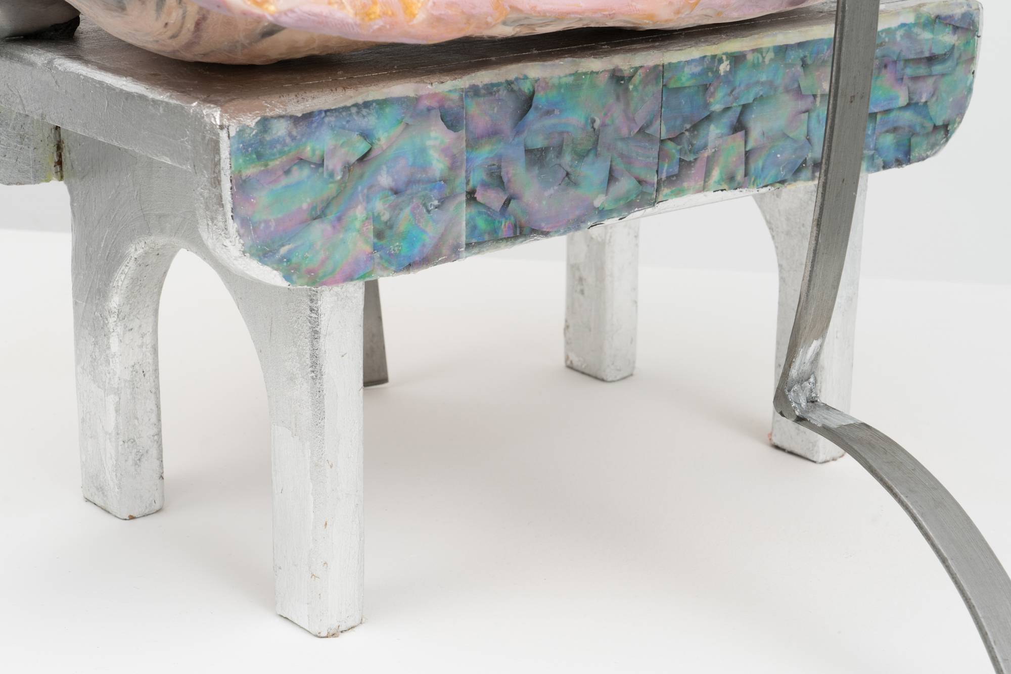 Detail of an Em Rooney sculpture featuring a stool wrapped in foil and mother of pearl, supporting a hinged steel mold of a flower, a pink fiberglass mold of an arm and a thin green silicone cast. A thin strip of bent steel is draped over the sculpture.