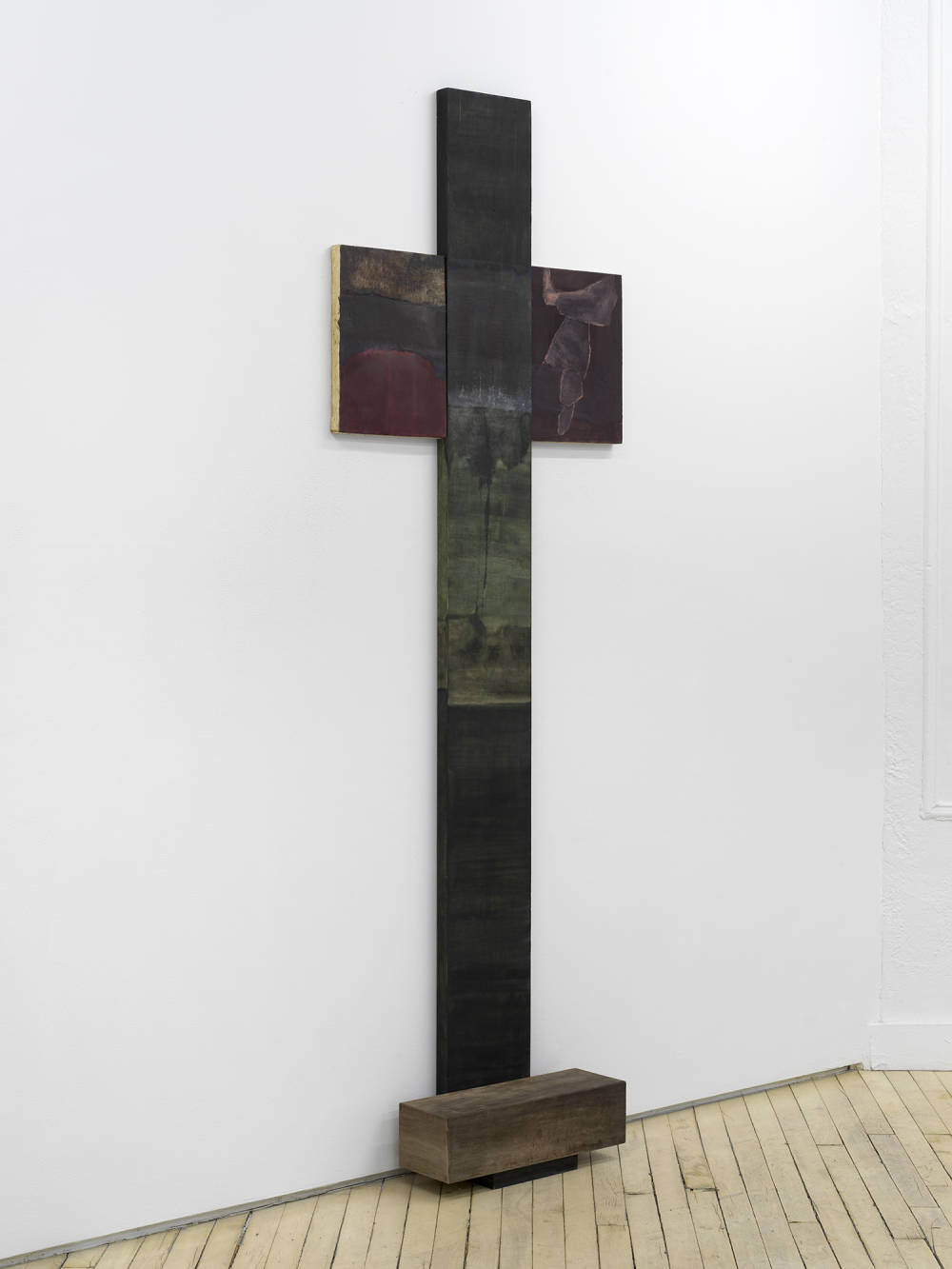 A vertical painting and sculpture in the shape of a cross leaning against a gallery wall. 