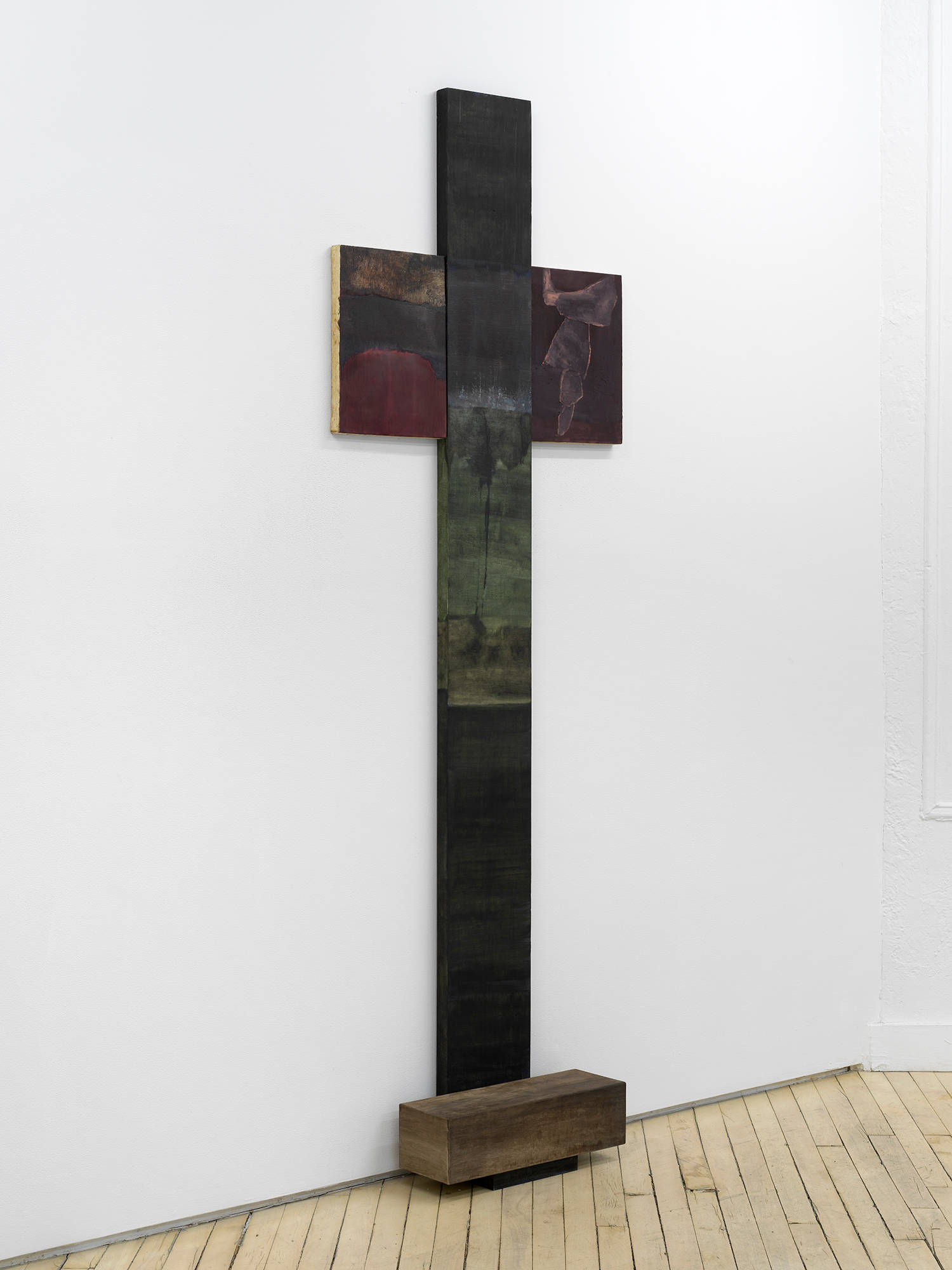 A vertical painting and sculpture in the shape of a cross leaning against a gallery wall.
