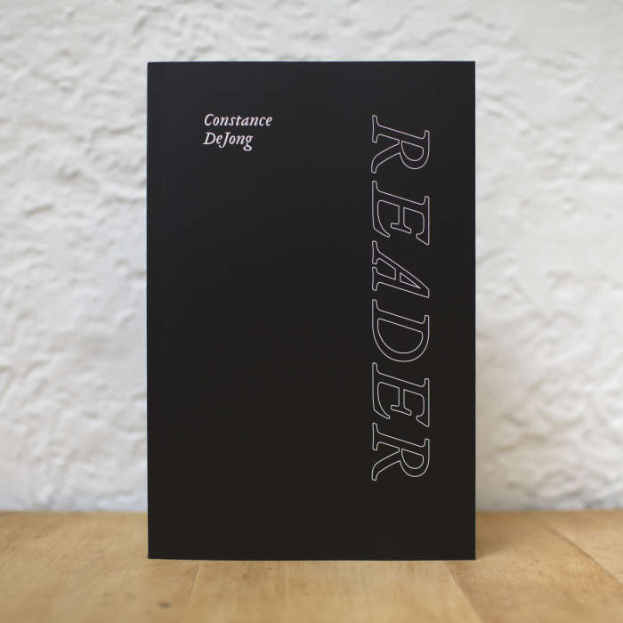 Image of Constance DeJong's new book, entitled Reader. The cover is black and white text and the book is standing on a table against a white background.