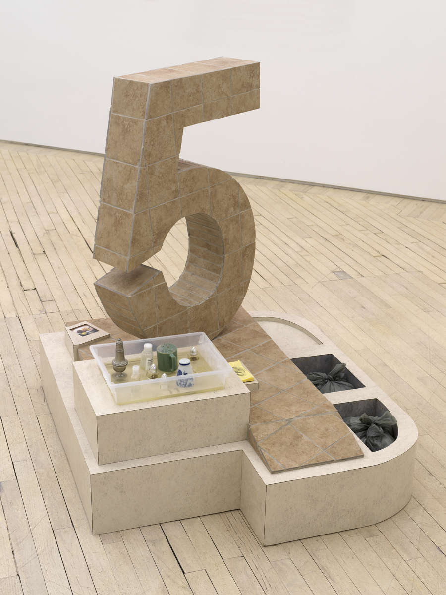 In a gallery space, a large abstract number 5 sculpture constructed out of flooring titles. The base of the sculpture contains two plastic bags and several salt and pepper shakers. There is a stack of post-it notes.