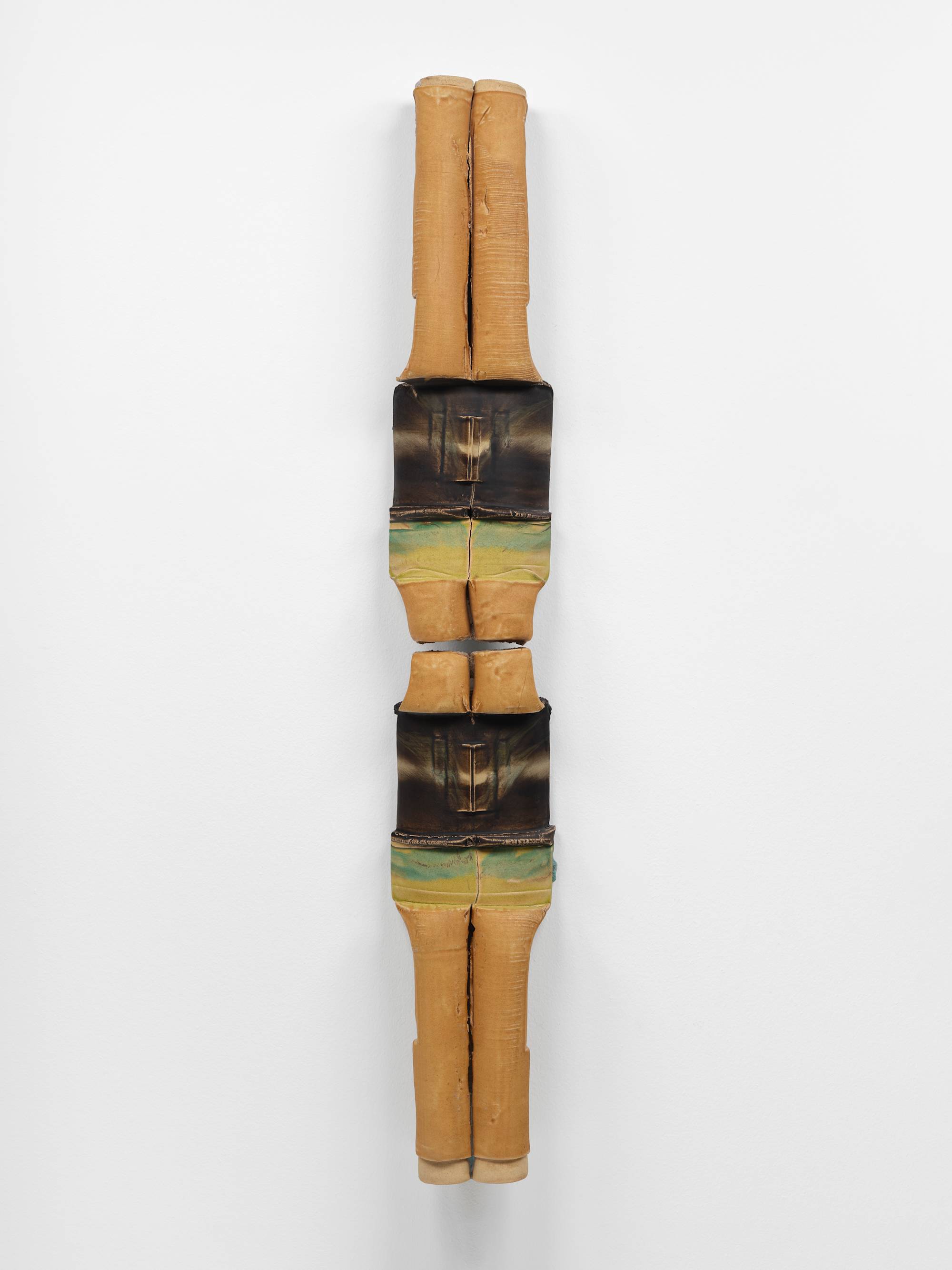 A large, vertically oriented, 2-piece sculpture, featuring cylindrical elements, in hues of mustard, brown, and green, hangs on a white wall.