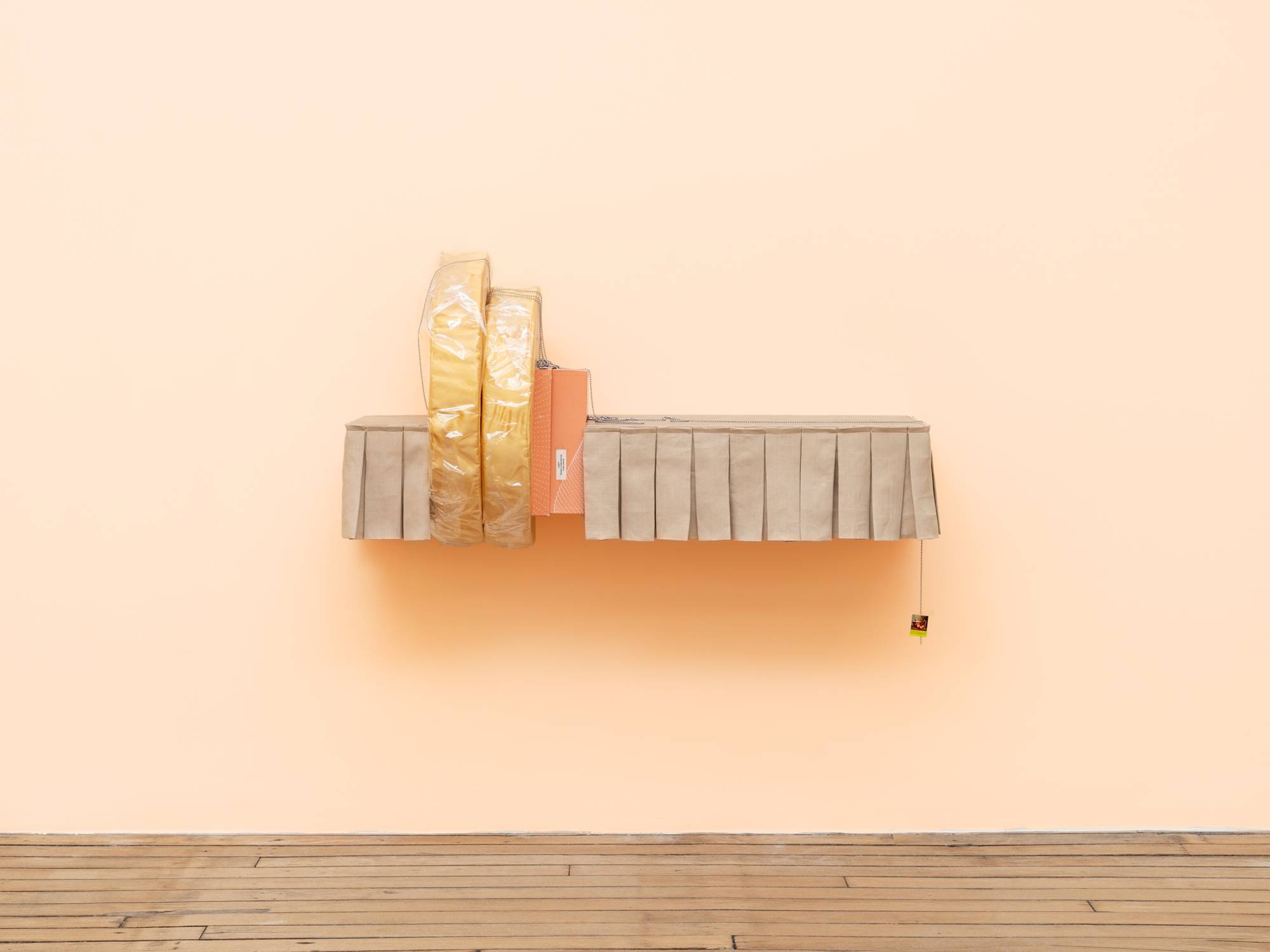 A sculptural shelf hangs on a peach colored wall. The shelf is lined with a short, beige, pleated curtain that is parted to the left of center by two vertical, yellow cushions, wrapped in cellophane, and an orange shoe box. A small picture hangs from the right side of the shelf by a ball chain.