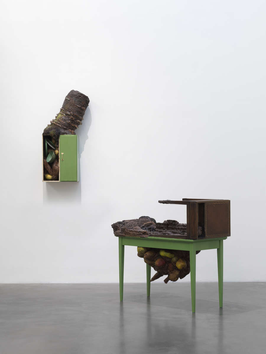 Two sculptures resembling constructed pieces of furniture painted green. Protruding out of both sculptures are tree-like, fungal forms. 