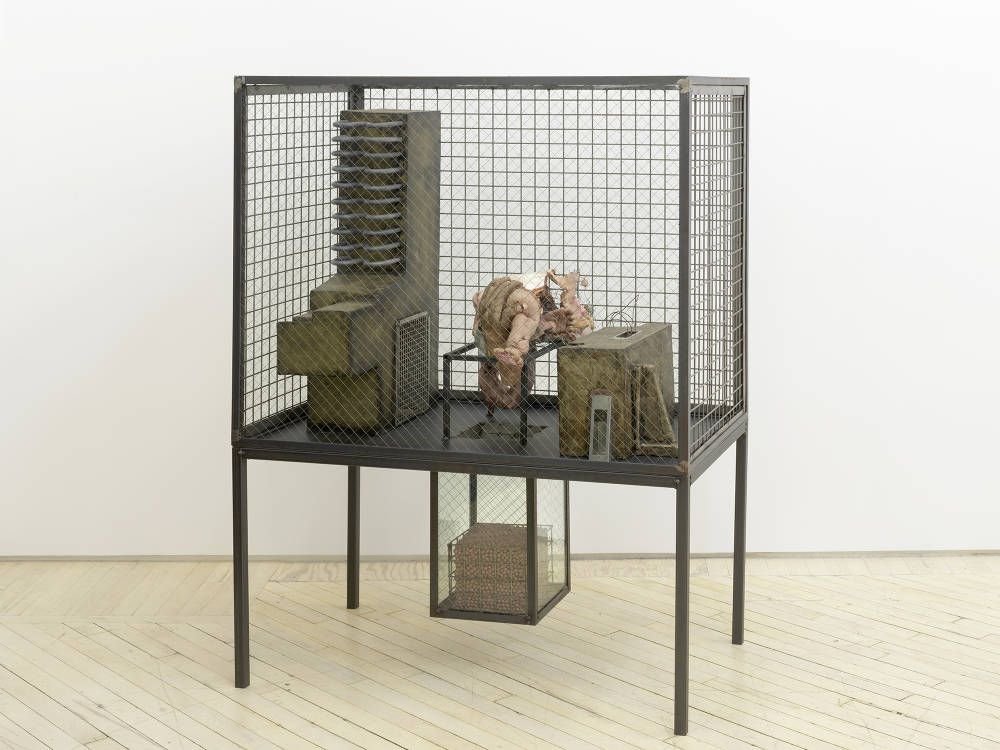 In a gallery space, a large iron sculpture resembling a cage containing architectural models and an abstracted body part.