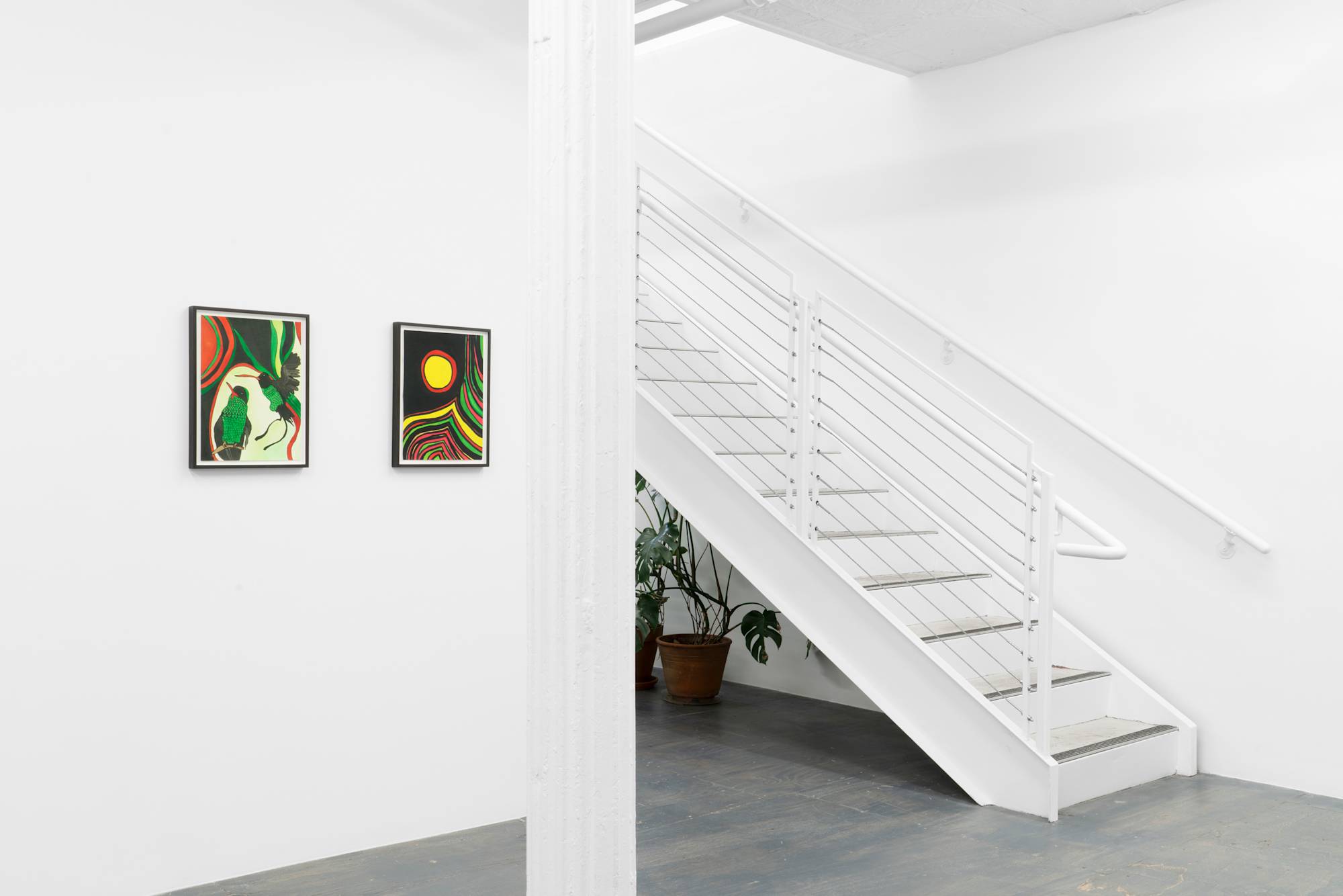 Two framed ink drawings hang on a wall to the left of a central column above a grey wood floor. A stair case ascends from right to left into the center of the frame. A potted monstera plant is visible under the staircase.