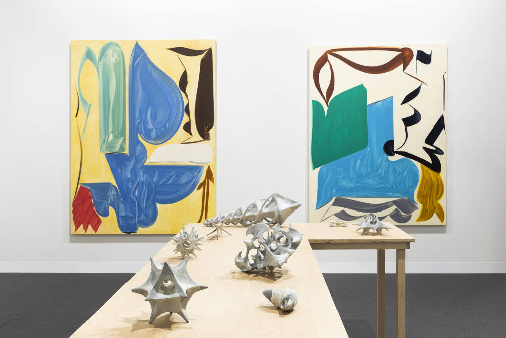 In a gallery space, two large abstract paintings are spaced apart. The painting depict bold abstract geometric shapes in various hues of color. The dominant colors are hues of yellow, blue, green, brown, and black. The paint is thin and gestural. In the center of the gallery space is a freestanding brownish table with various miniature gray sculptures placed evenly apart. The sculptures are abstract and resemble organic material or bone formations. 