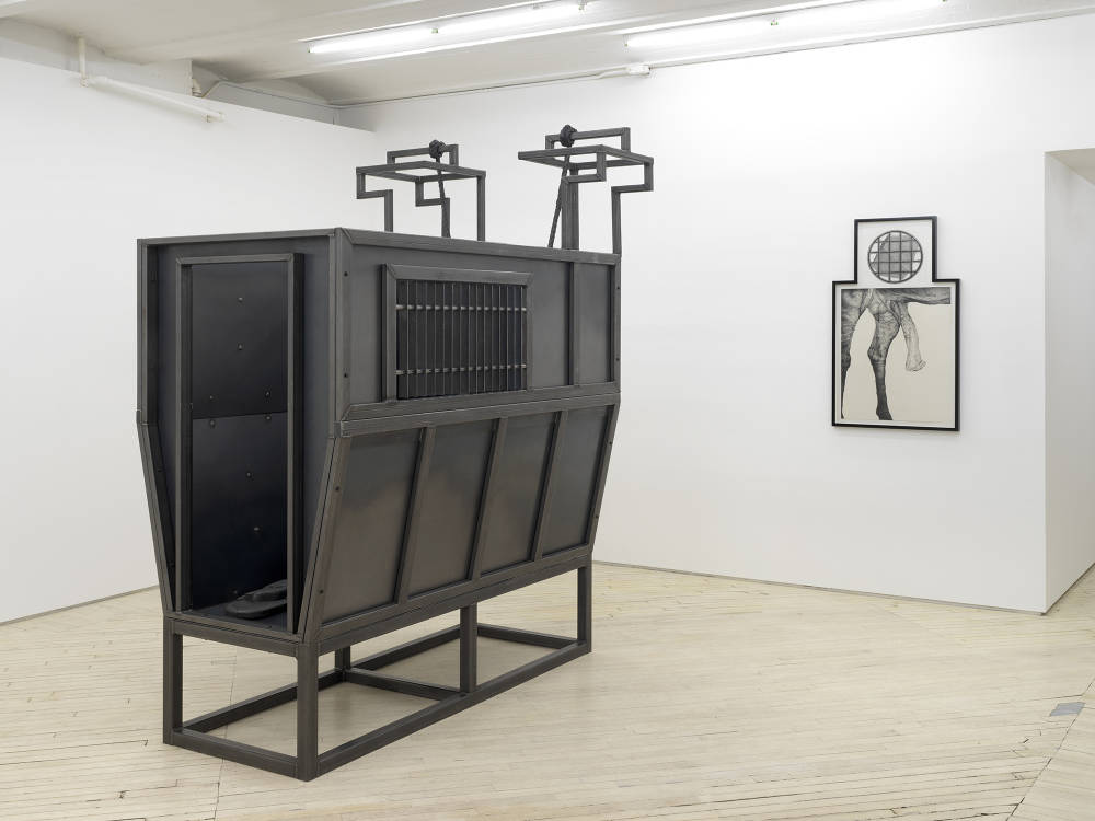 In a gallery space, a large floor iron floor sculpture resembling a cattle container. Behind the sculpture hung on the wall is a graphite drawing of horse genitals with a geometric pattern in a circle drawn above it in an irregularly shaped black frame. 