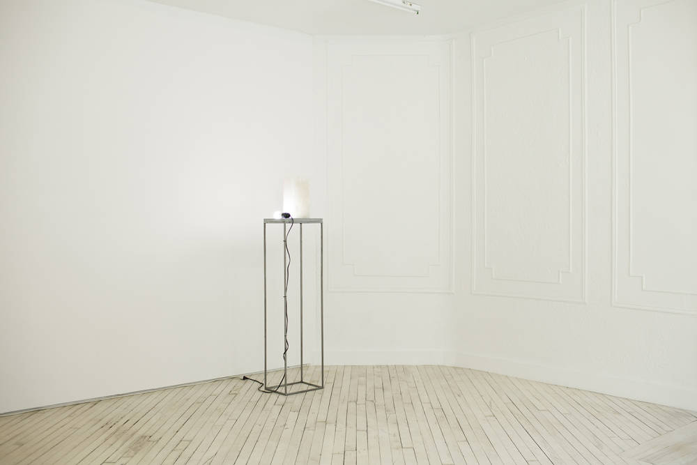 In the corner of a white gallery is a tall steel and stone plinth with another rectangular block of alabaster sitting on top, there is an illuminated light bulb also sitting on the plinth with its wire running down to the floor.