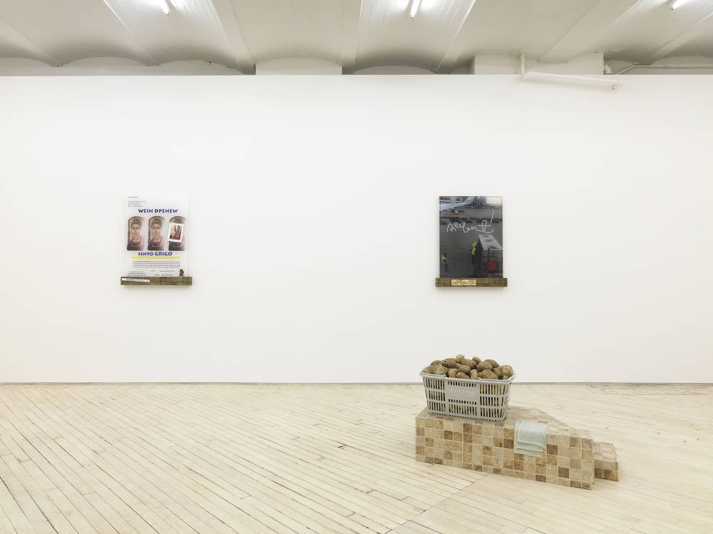 In a gallery space, two wall sculptures are hung evenly spaced apart on a large white wall. They contain printed images on plastic. One the photographs is of an airport. The base of the sculpture resembles a row of bricks. On the floor is a sculpture constructed out of floor tile. Resting on top is a shopping basket full of potatoes. 