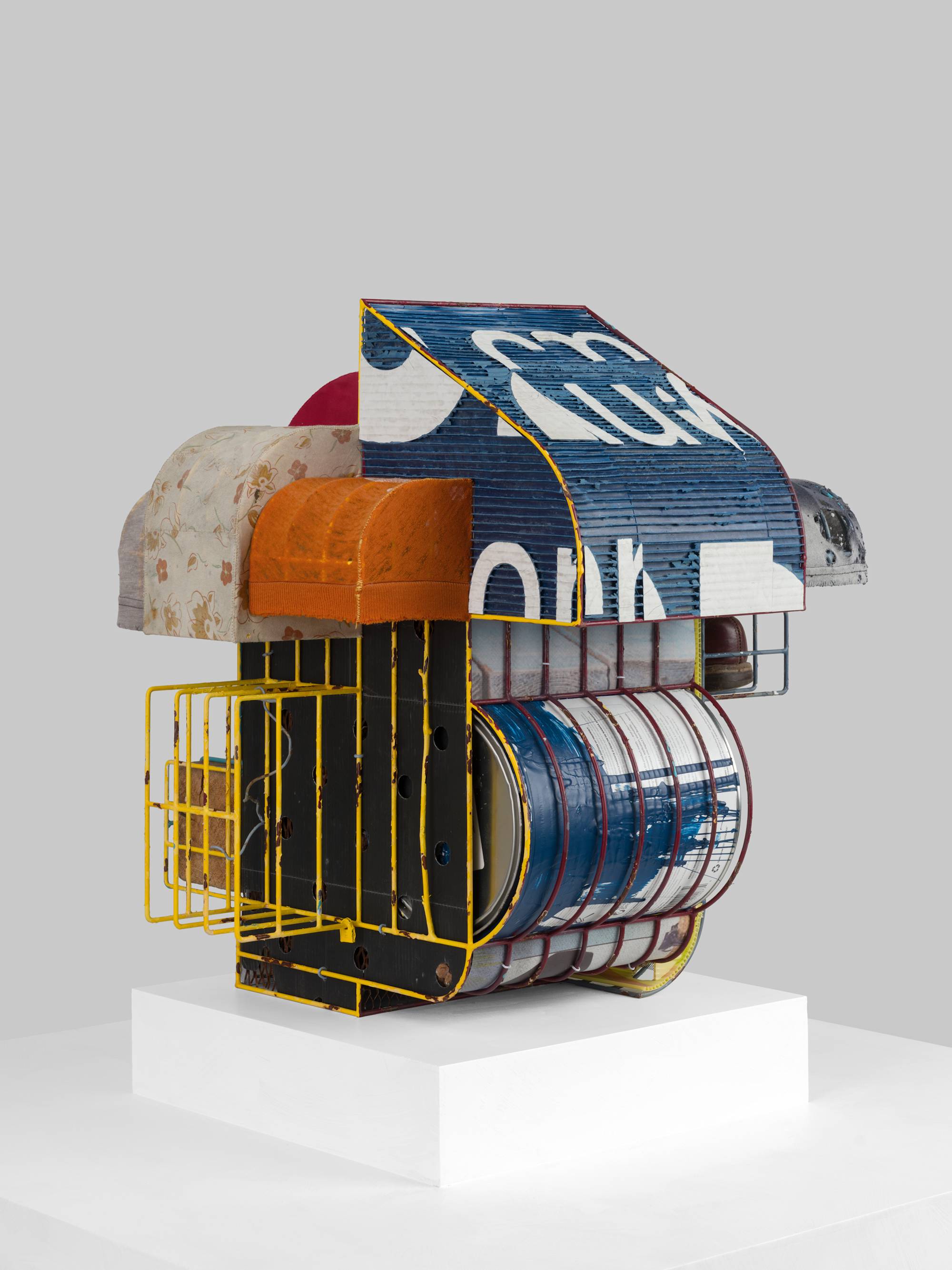 A cage-like colorful steal wire sculpture, wrapped with several tattered fabrics to resemble various awnings, and embellished with an old shoe, a twig, printed coroplast signage, an old blue paint can, a lock, a paper tag, and a wooden block sits on a white pedestal against a white wall.