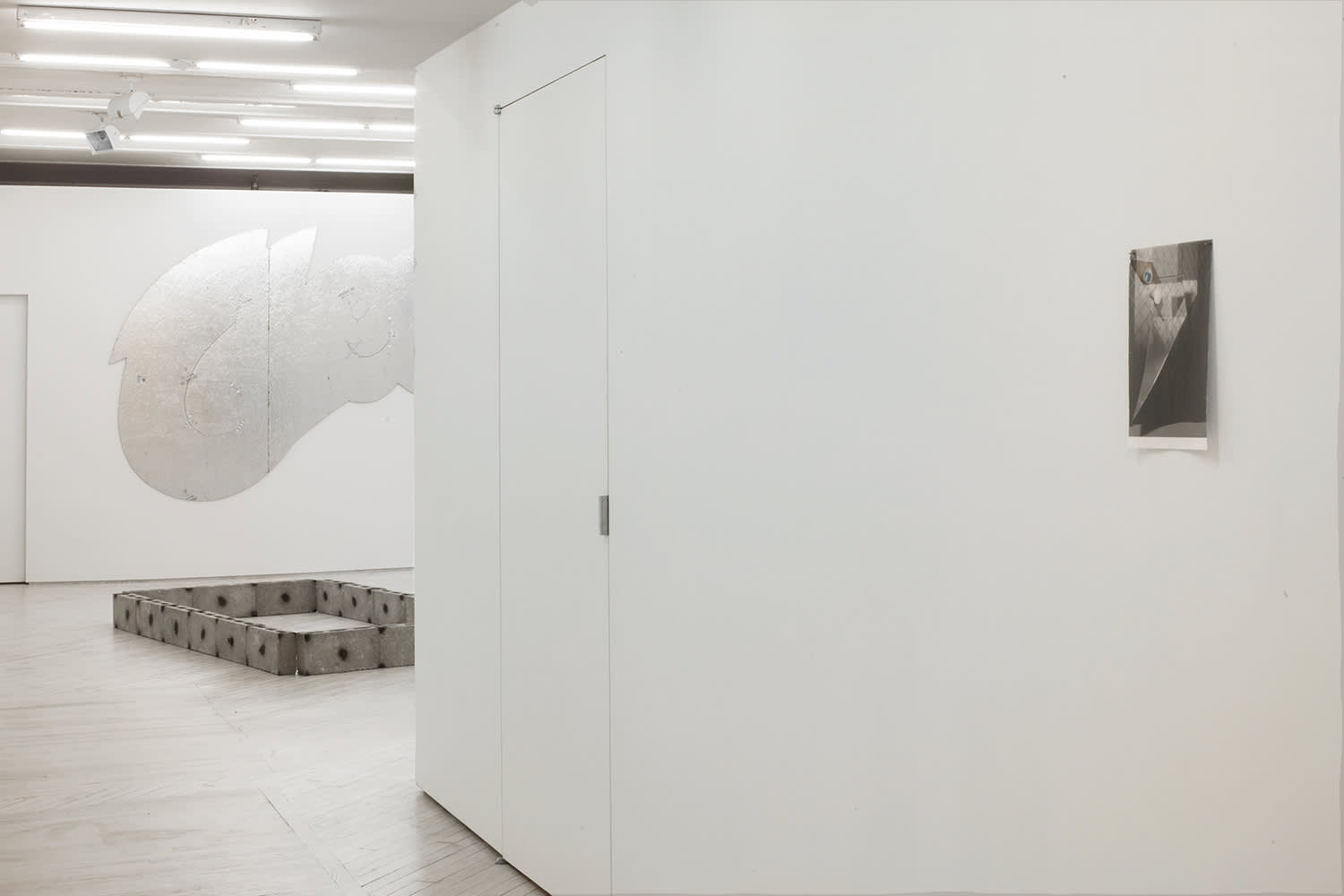Installation view of Piss Yellow Bars and Stars, a solo show by Tom Holmes from 2013. View down the hall, with a small fabric work in greyscale on the right wall. One floor sculpture made of grey cinder blocks in the foreground - adorned with black spray paint dots. A shaped silver foil cut out painting in the shape of the Cheeto's logo hangs on the back wall, from behind the hallway wall.