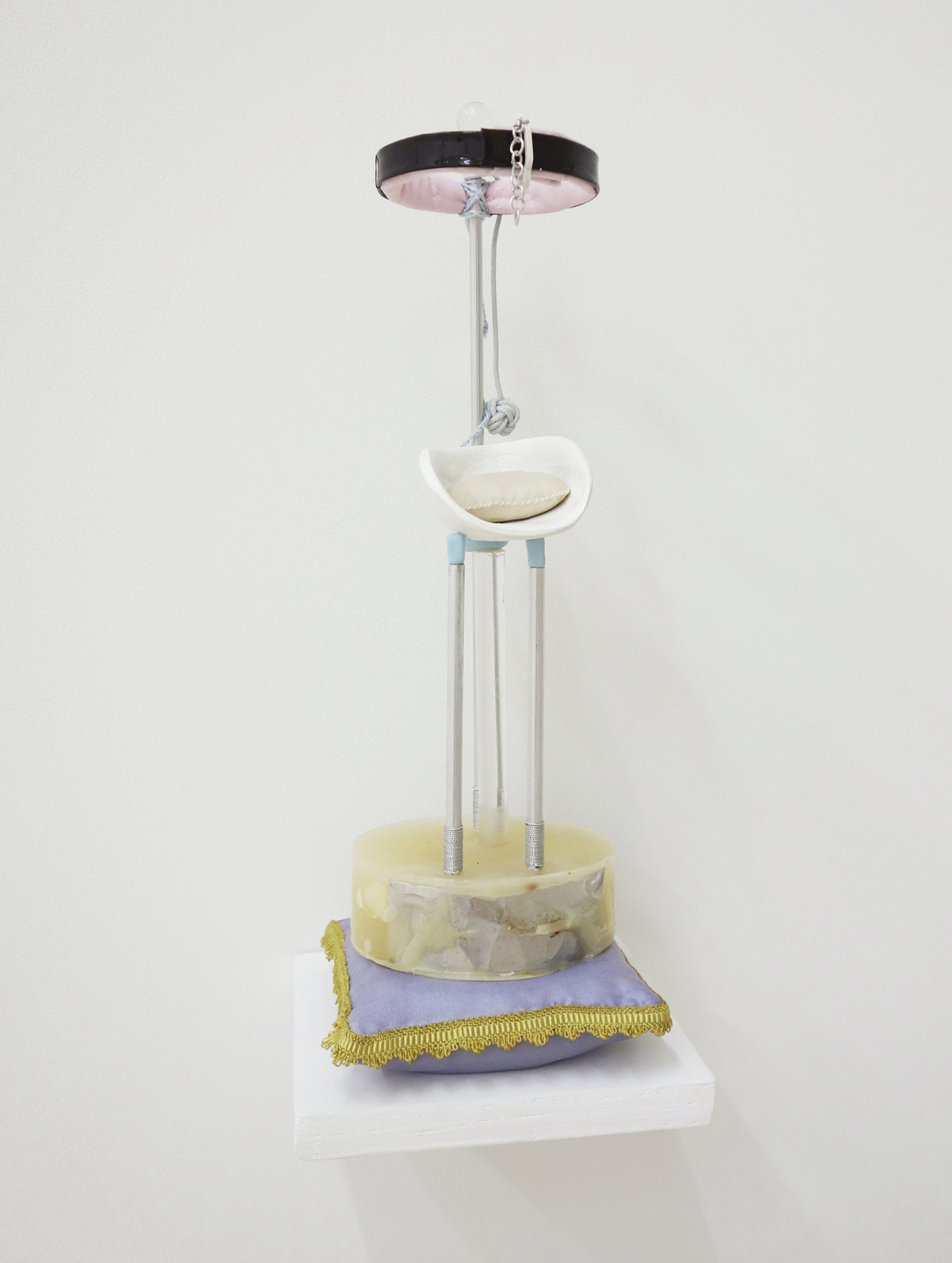 Small sculpture made of a small purple pillow with yellow trim and a small, tiered, tower-like contraption on top of it, the center features a hand made tiny white pillow cradled in something rounded and the top has a black collar type object lined with pink satin.