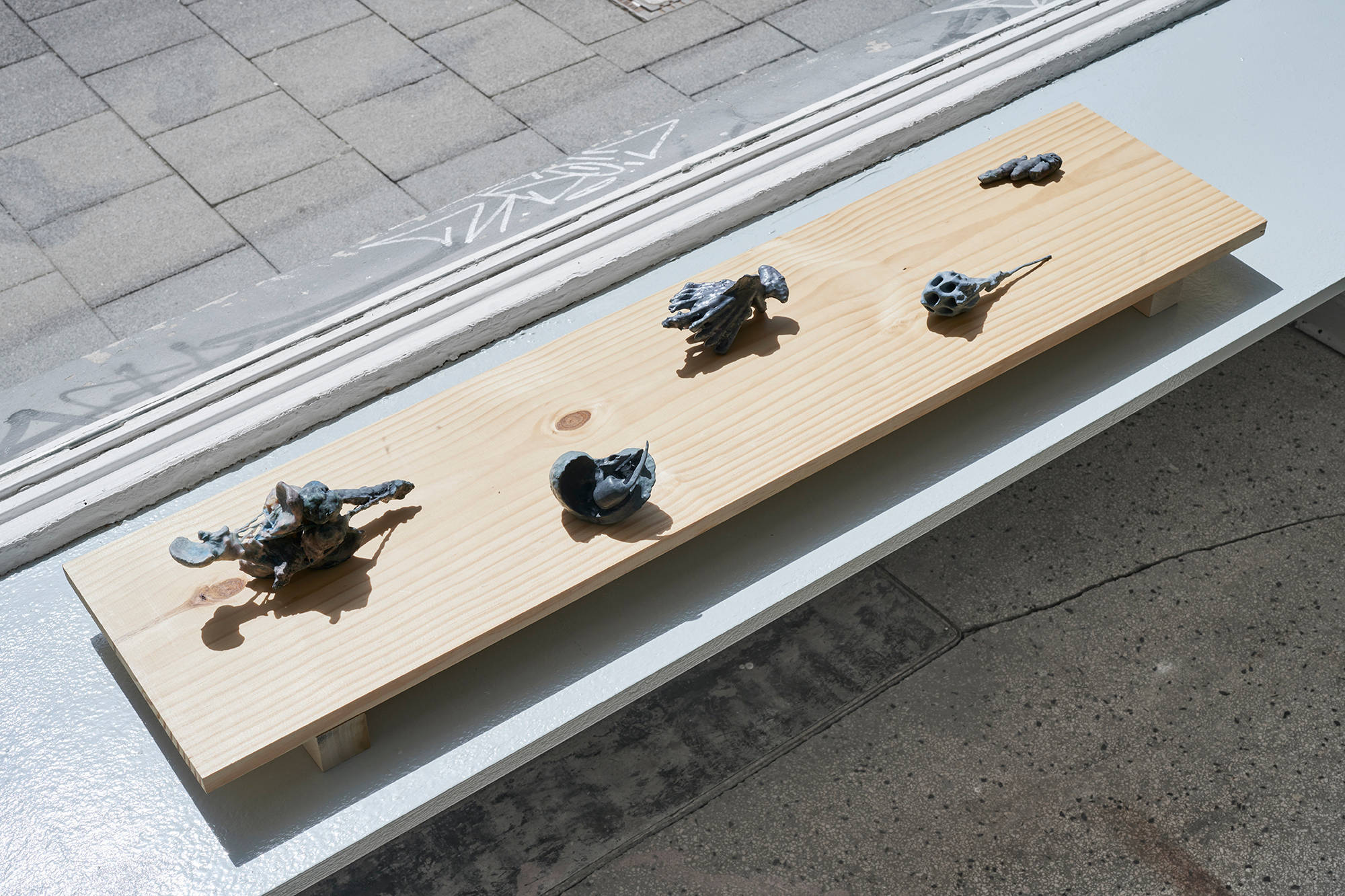 Five small hand-sized sculptures are presented on a rectangular piece of wood. The sculptures are amorphous, skeletal, and gray.