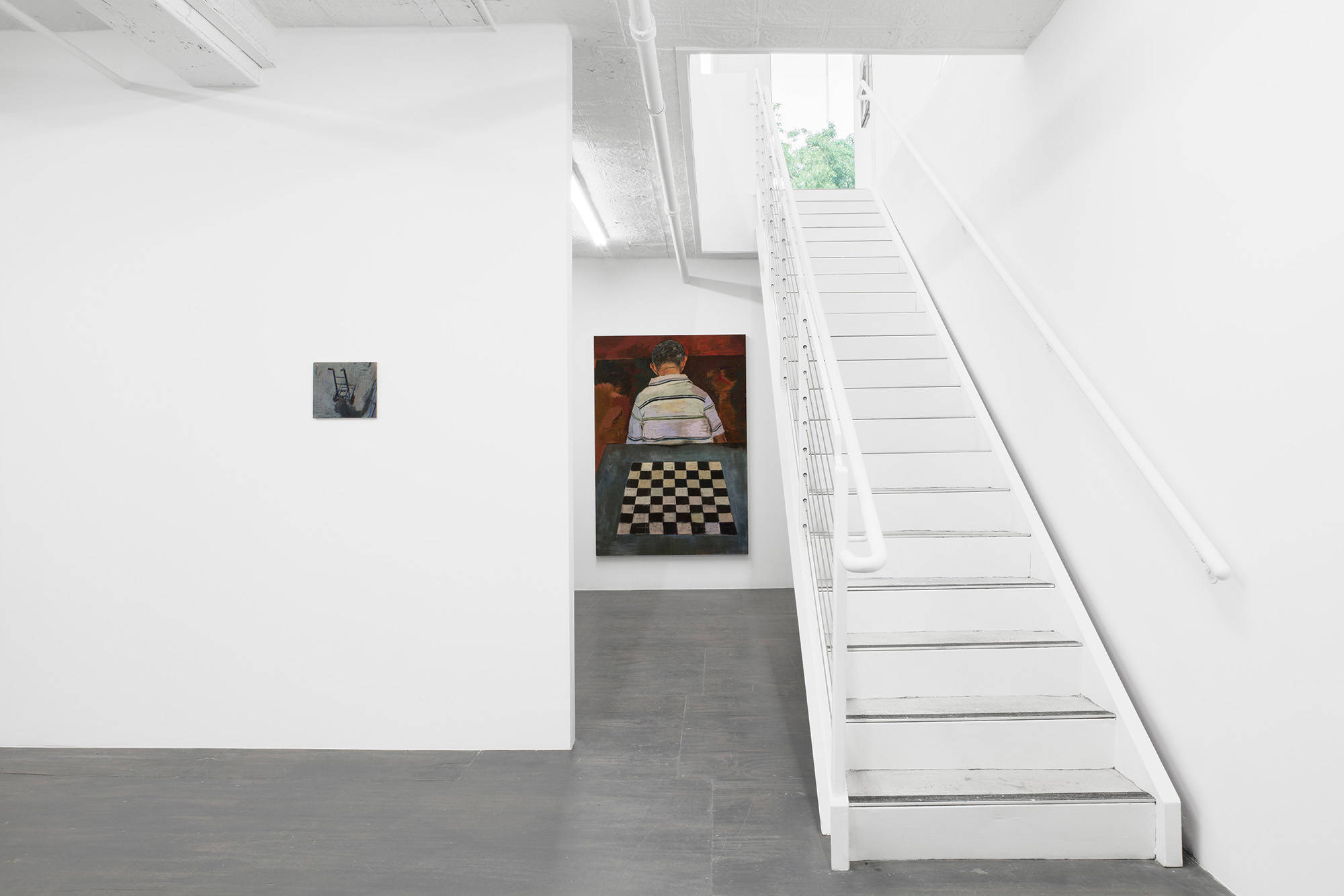 In this view of the North wall in the downstairs of the gallery, there is a stairwell to the right of frame, extending to an upper floor. To the left of frame a wall holds a small painting of a dolly. Between the stairs and the wall, a painting of a checker board and a figure in a shirt can be seen hanging on a further wall.