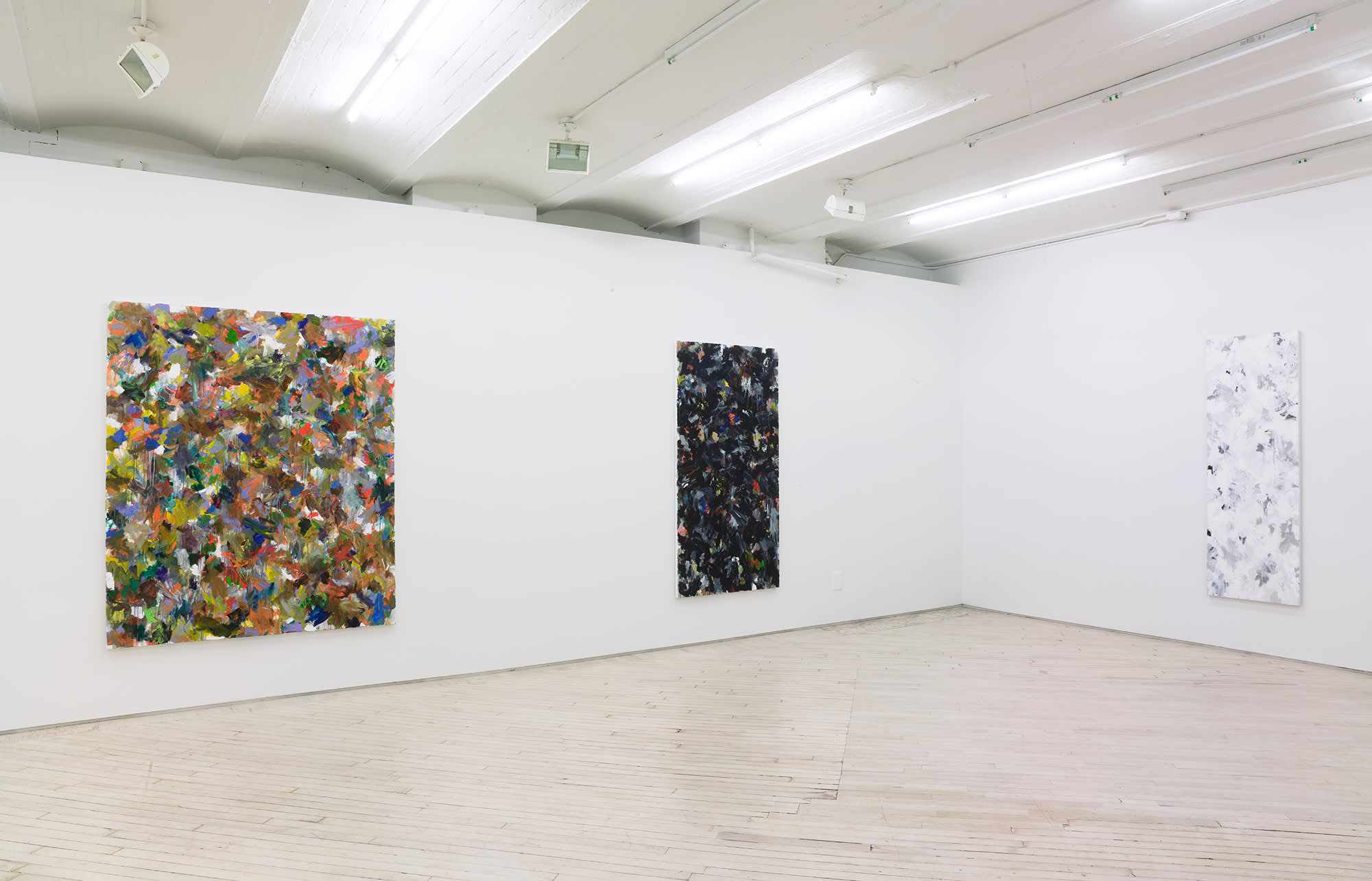 In a gallery space, three large abstract paintings are hung evenly spaced apart on large white walls. The paintings contain numerous swatches of various colors painted on top of one another. The paint is thin and in some areas dripping.