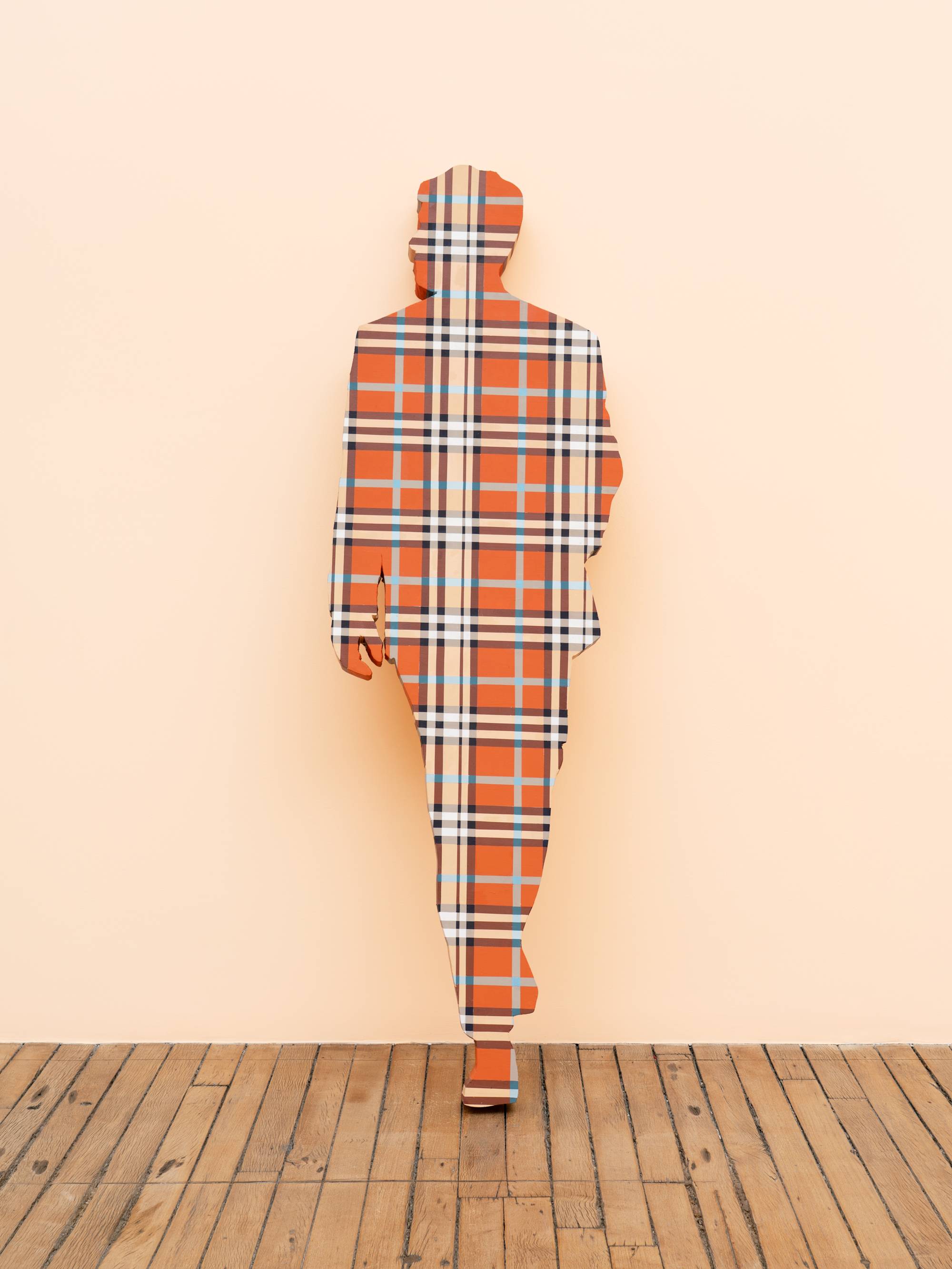 A life-sized human figure painted in a blue and orange plaid pattern leans against a peach wall in an art gallery.