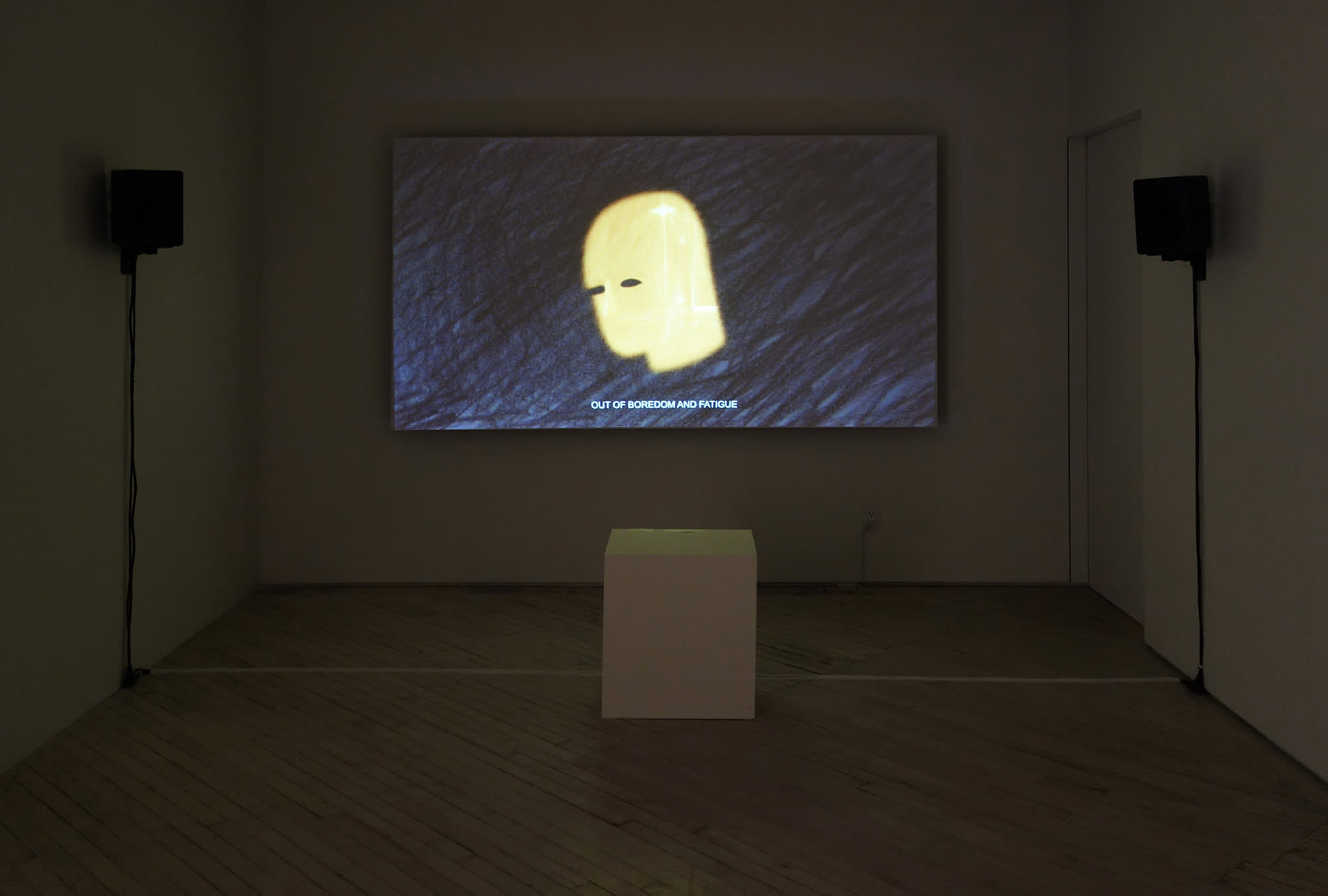 Installation image of a video room with two speakers to left and right and a color 6:9 image at center, at bottom a white cube seat. The video image is from a hand drawn animation. Dark grey background with grey and black sketchy marks of pencil, at center is the yellow silouhette with black eyes of a man's head in 3/4 profile. At bottom is a subtitle which reads, "OUT OF BOREDOM AND FATIGUE".