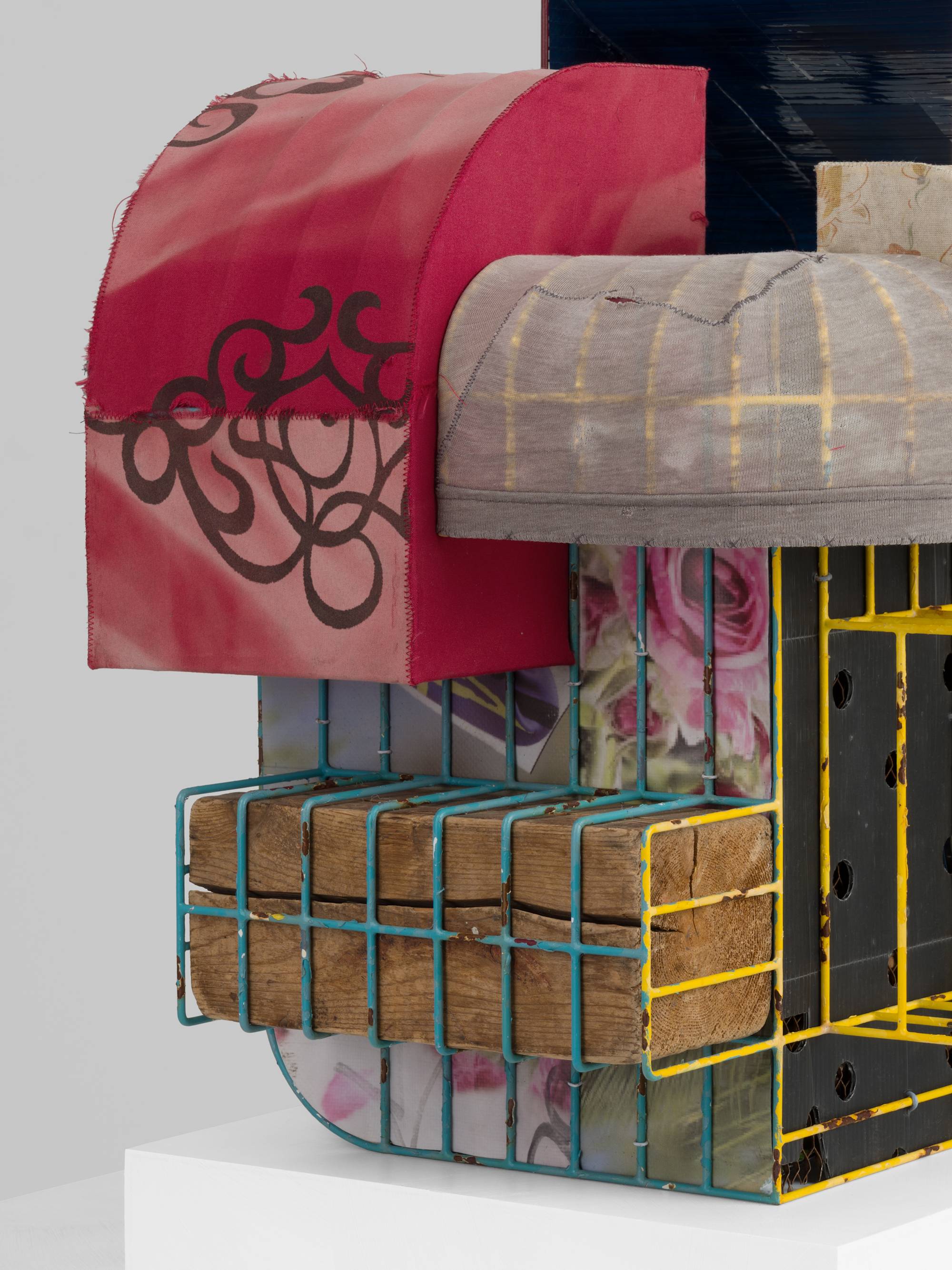 Detail of a cage-like colorful steal wire sculpture, wrapped with several tattered fabrics to resemble various awnings, and embellished with an old shoe, a twig, printed coroplast signage, an old blue paint can, a lock, a paper tag, and a wooden block sits on a white pedestal against a white wall.