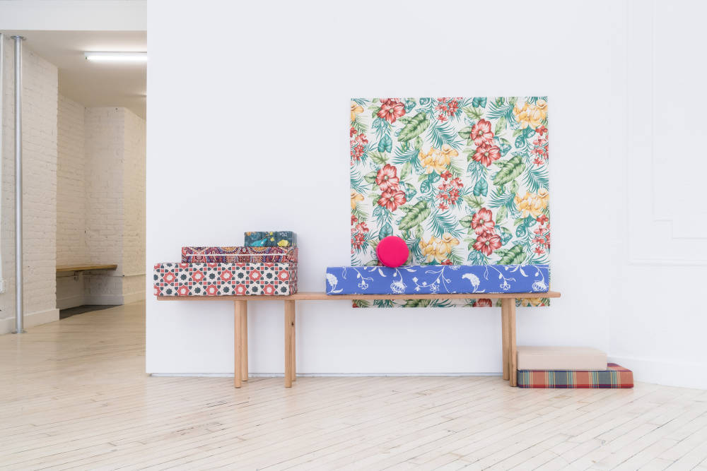 Installation view of Jaya Howey exhibition at Bureau, New York with a custom designed fabric swatch pinned to the wall behind a six legged long wooden bench with an array of rectangular pillows on top and on the floor.