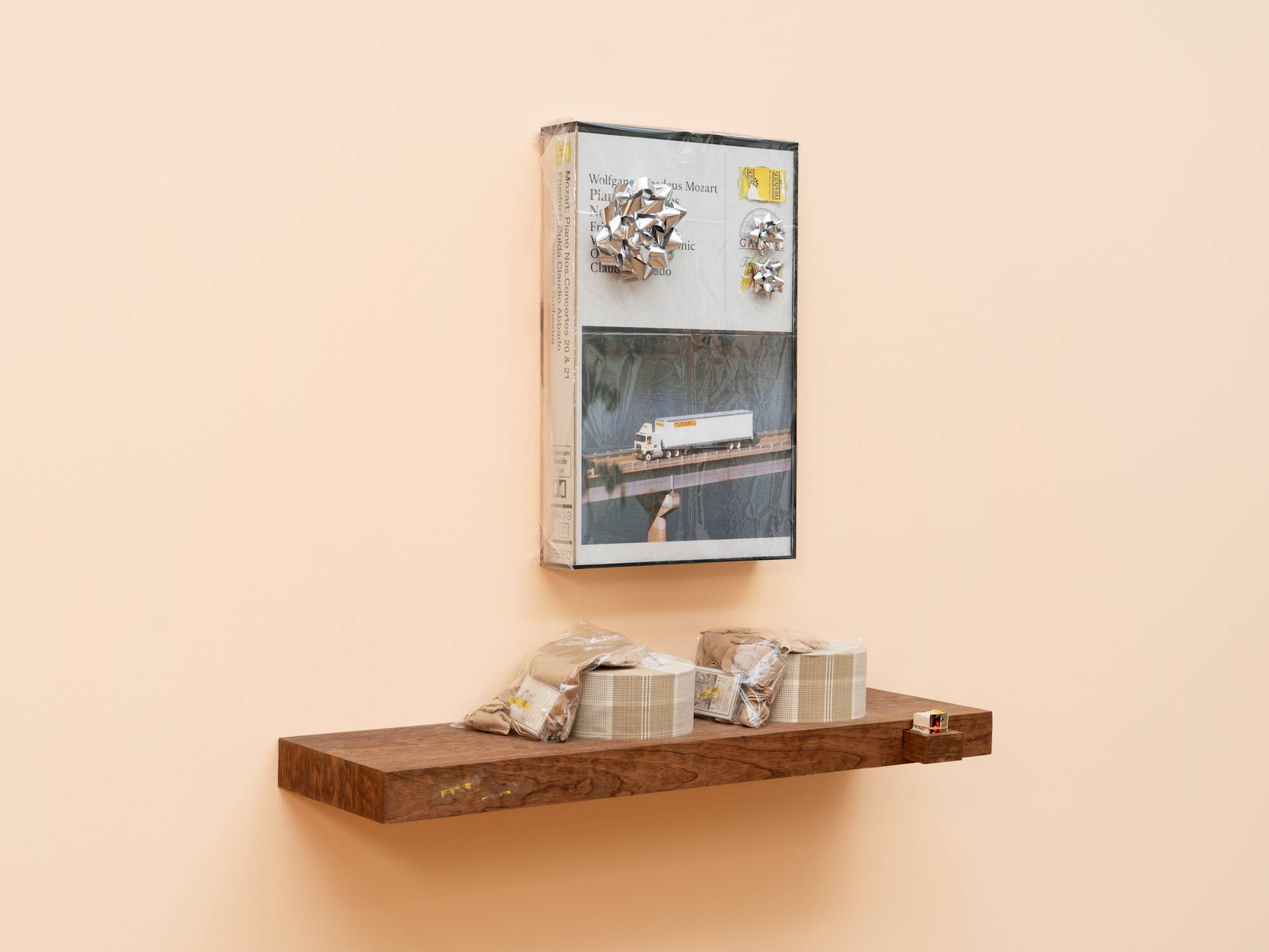 Angled view of a wood veneered shelf hangs on a peach colored wall. 2 tan, plaid, circular gift boxes sit on the shelf with tan socks in plastic bags draped over them. A small drawer juts out of the front of the shelf and is neatly filled with trimmed business cards. A large, fake audio cassette case featuring a truck driving over a bridge hangs above the shelf, wrapped in cellophane and decorated with three silver gift bows.