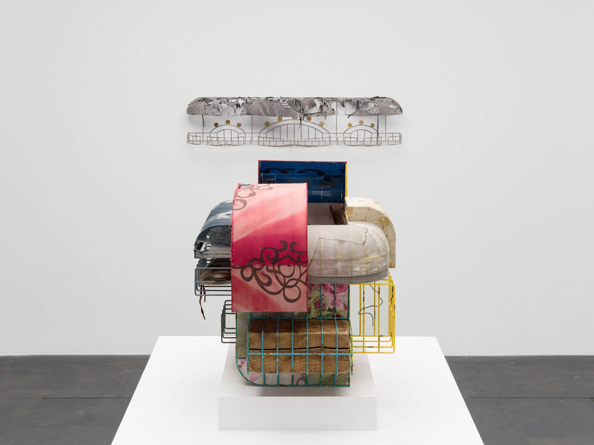Straight on view of a colorful, cage-like sculpture, wrapped in tattered clothing, standing on a pedestal in the center of a white walled gallery with grey floor. Behind the sculpture another sculpture, with similar features, can be seen mounted to the wall.