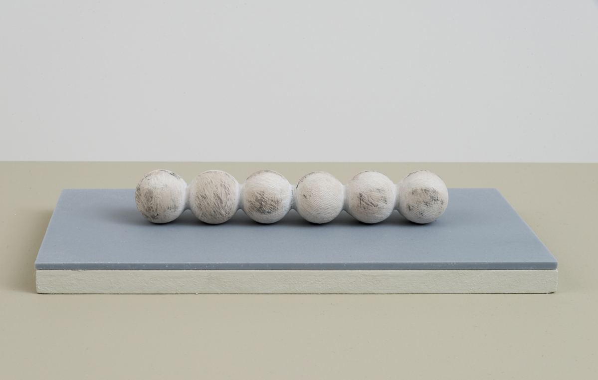 A handmade sculpture resembling a connected row of round bulbs resting on top of a blue-tinted plinth with a white wall partially in view. 