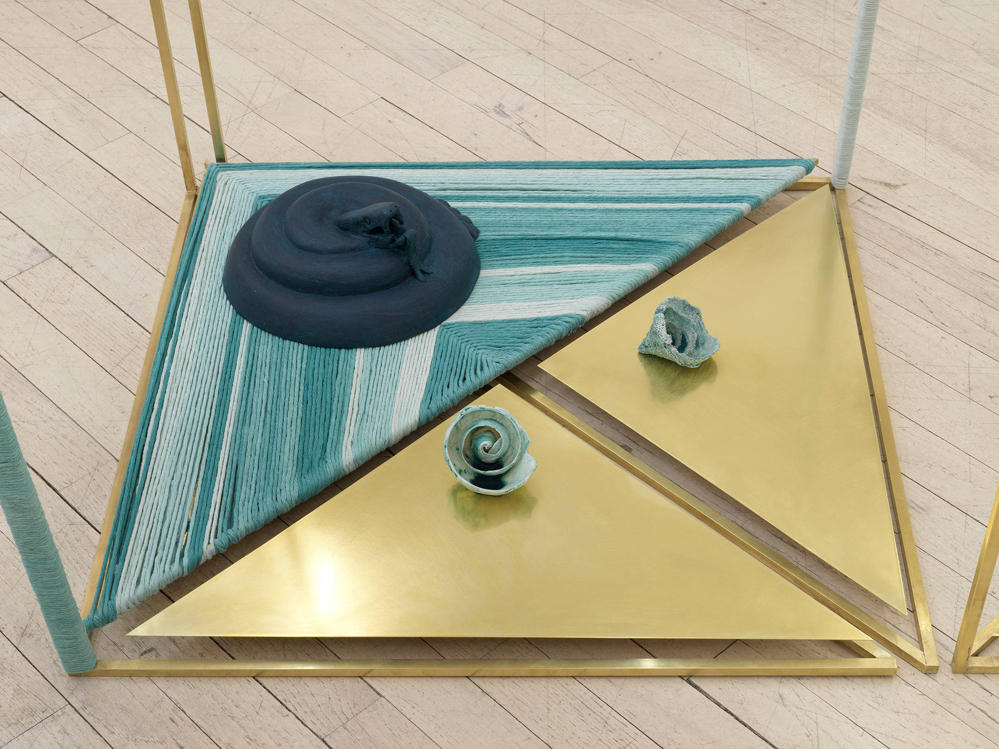 Image of a sculpture by Claudia Peña Salinas composed of vertical brass rods creating rectangular shapes in space with parts of the rod wrapped in dyed blue fabric. Sitting on the base of these rectangles are small sculptures of Mayan symbols.