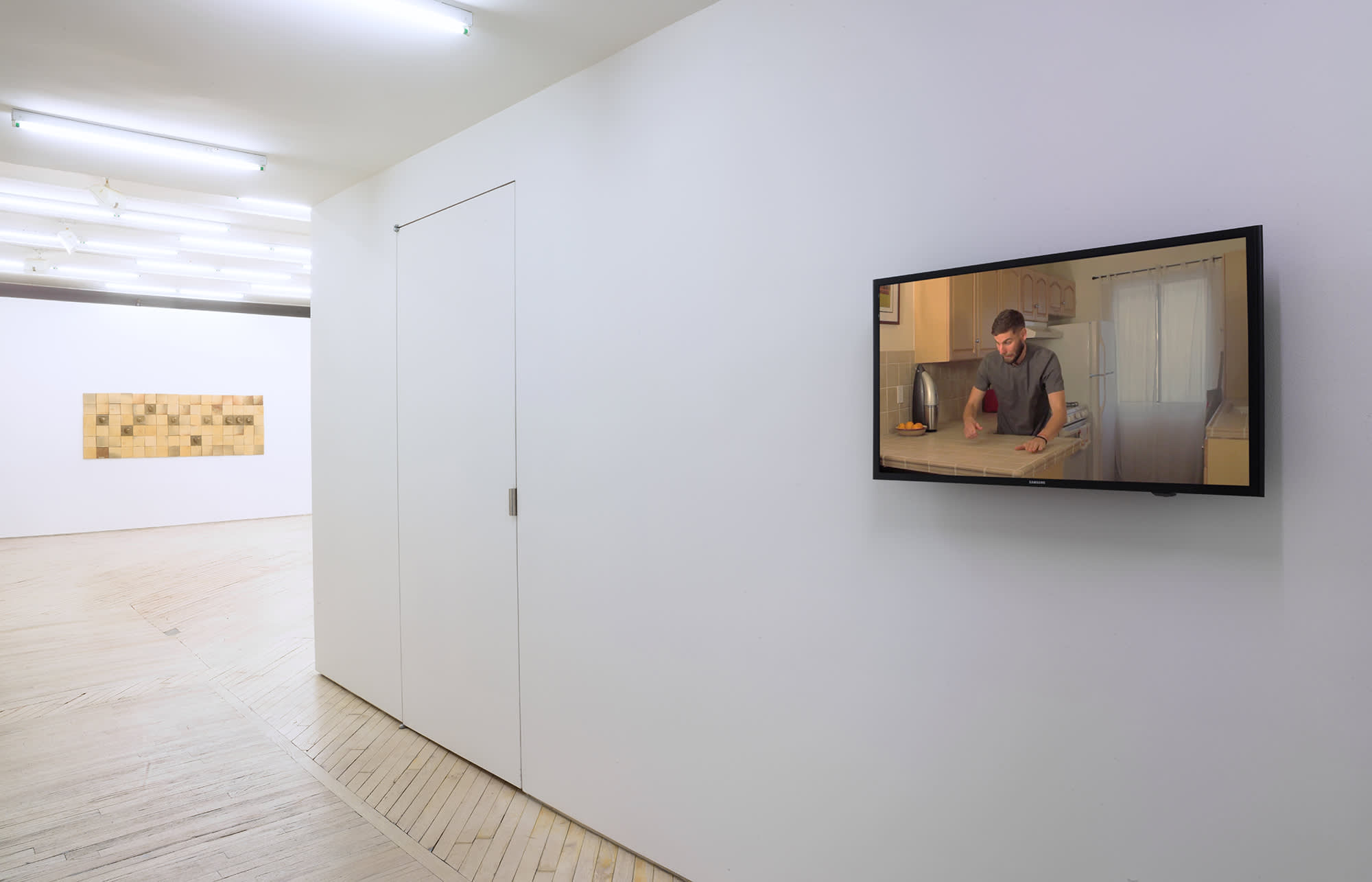 A view looking down a white hallway with the larger gallery at the end, a video screen hangs at right on the wall, in the video, a white young man is in a kitchen, with his hands on the countertop, looking down.
