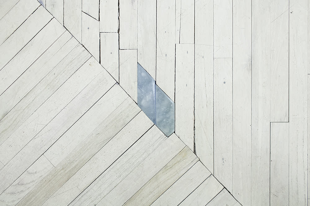 Image of the floor - white washed wooden floor boards, laid out at an angle, meeting at a diagonal across the photo, with a small trapezoidal shape removed along the center, which has been replaced by alabaster which has been tinted with light blue watercolor.