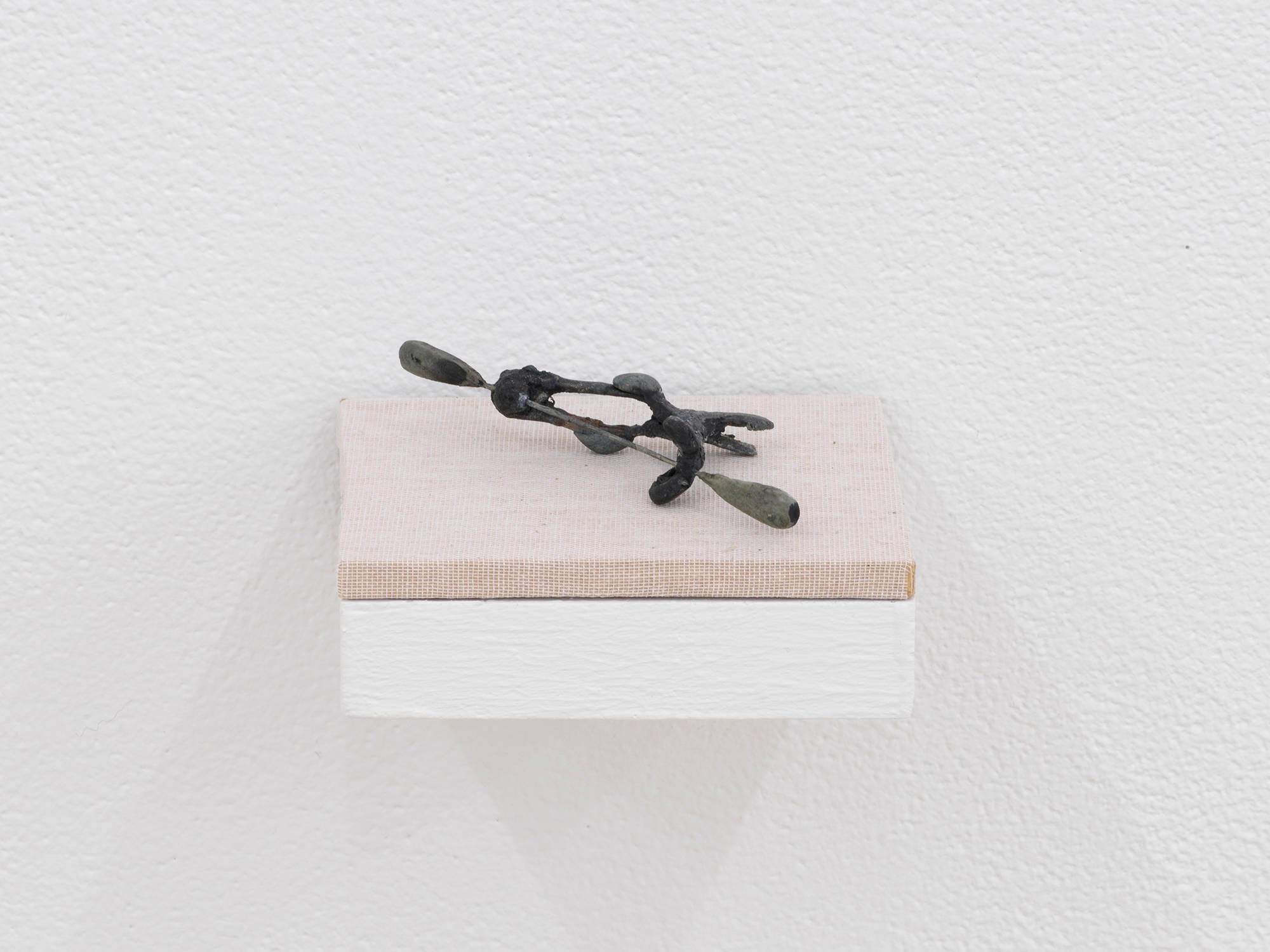 A small abstract sculpture resembling a fossil, bone formation, or combination of organic materials. The object is primarily gray and resting on a shelf with a tan base attached to a white wall.