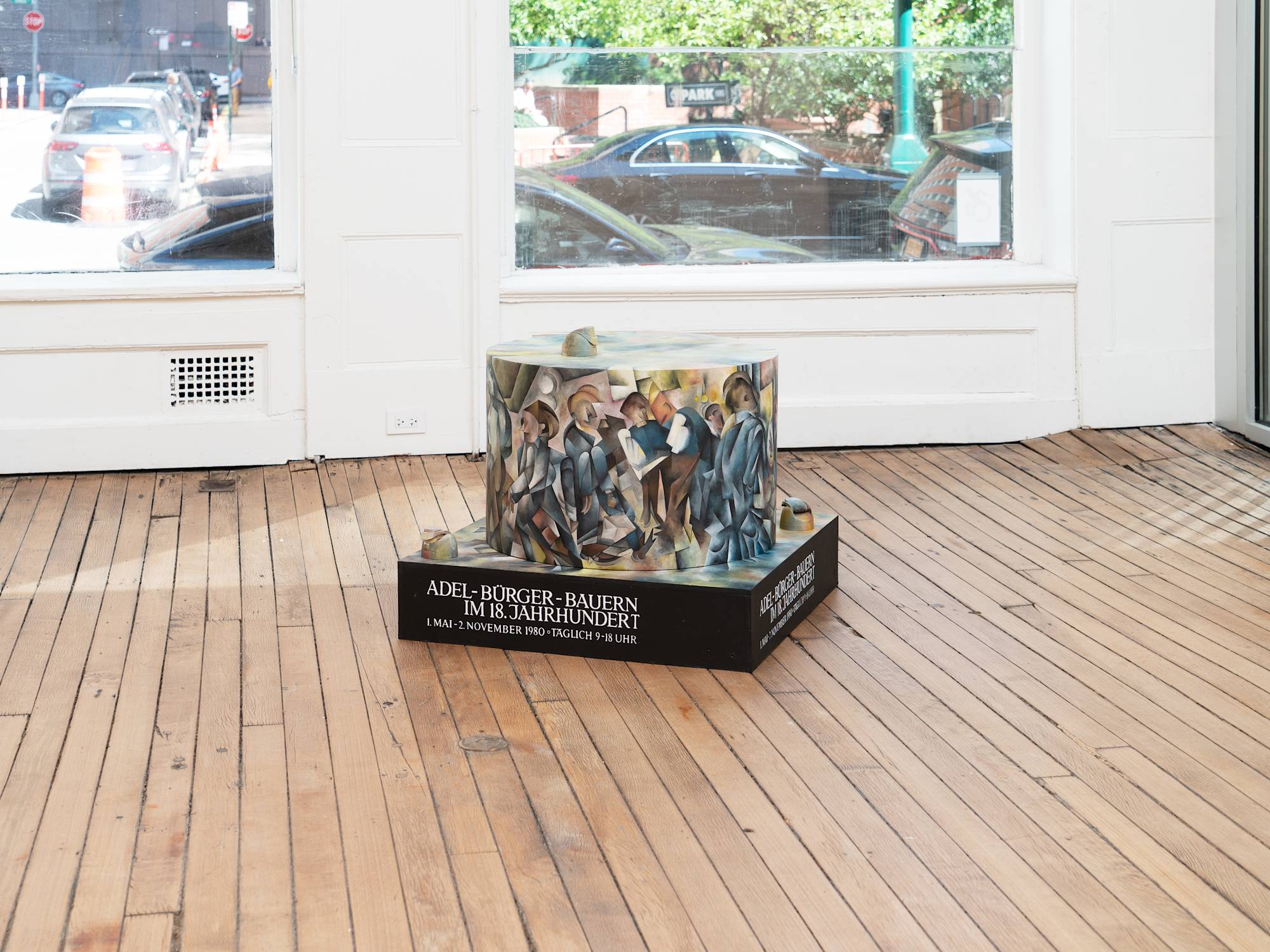 A freestanding sculpture with a round center sits in a gallery space in front of a window. The sculpture is painted in a Cubist style.