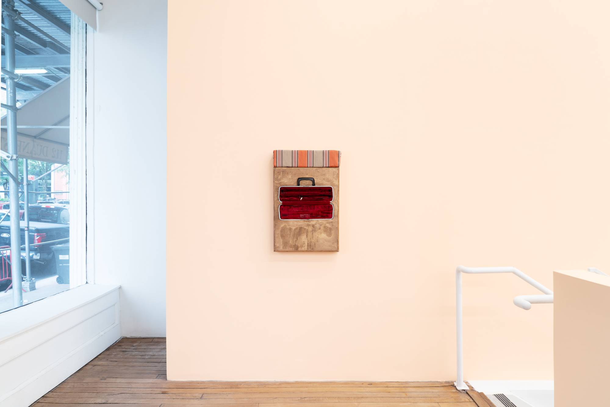 Installation view of a wooden box sculpture with inset flute case with red velvet lining, topped with a orange and beige striped cushion, hanging on a peach colored wall.