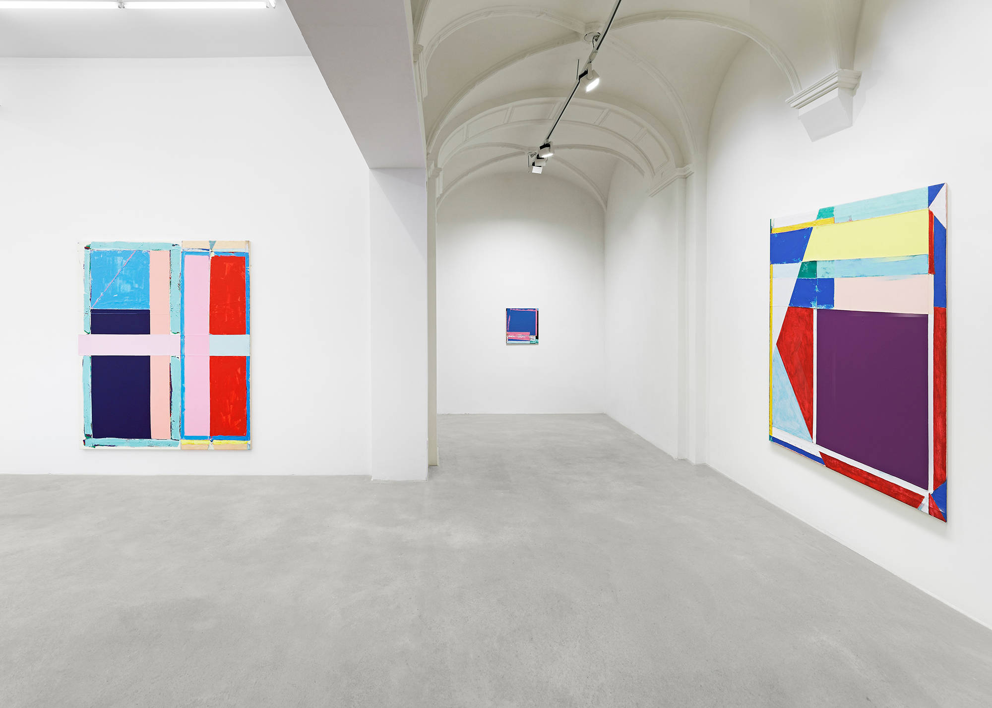 In a gallery space, two large abstract paintings hung on separate walls. The paintings are formally dominated by large rectangular blocks of color. The most striking colors in both paintings are a range of reds, pinks, blues, and yellows. The paint is thick and the canvases are rough. In the background is a smaller blue and pink painting.