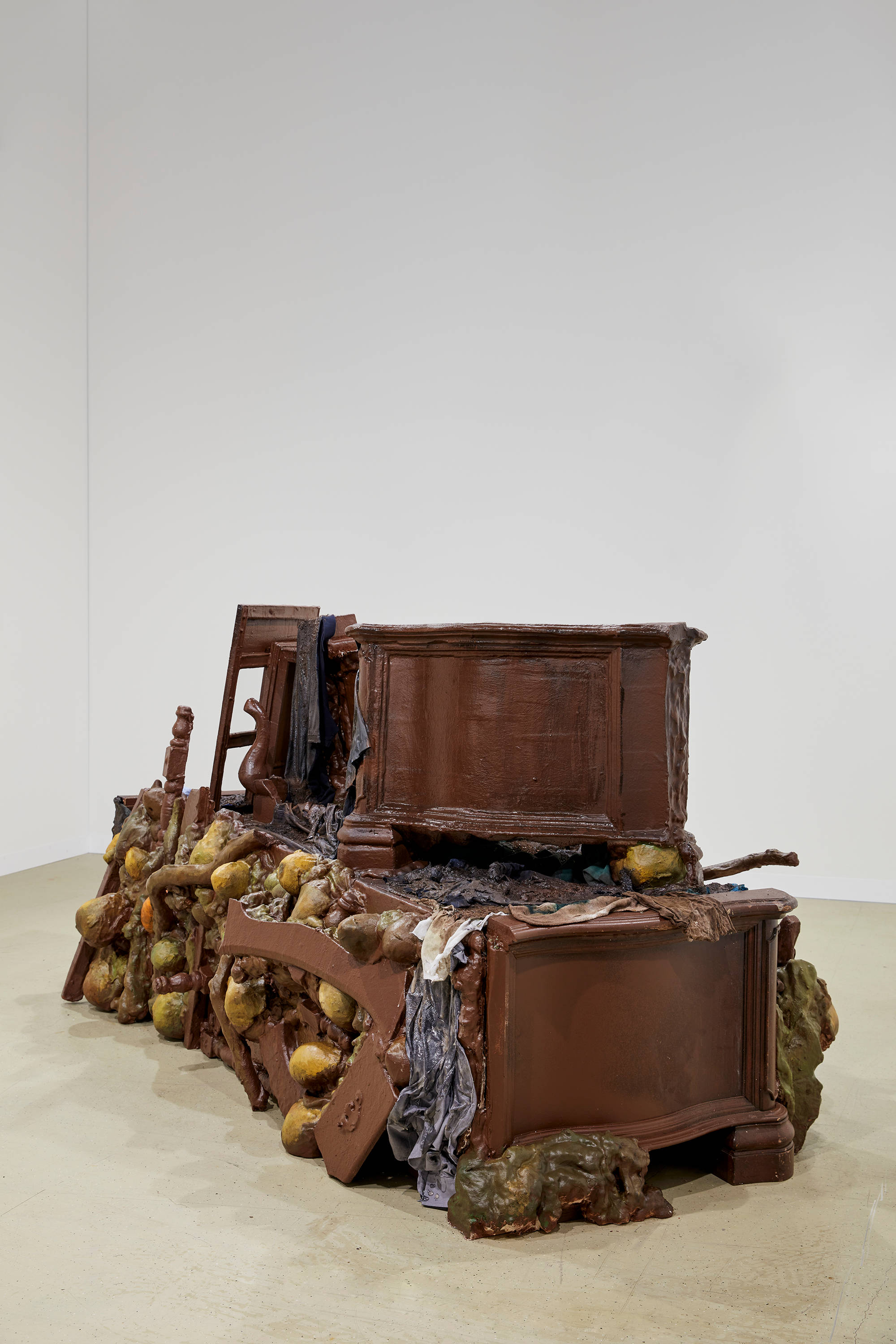 A large piece of brown furniture sits in the middle of a room with white walls. A smaller sculpture hangs to the right of the larger installation on the wall.