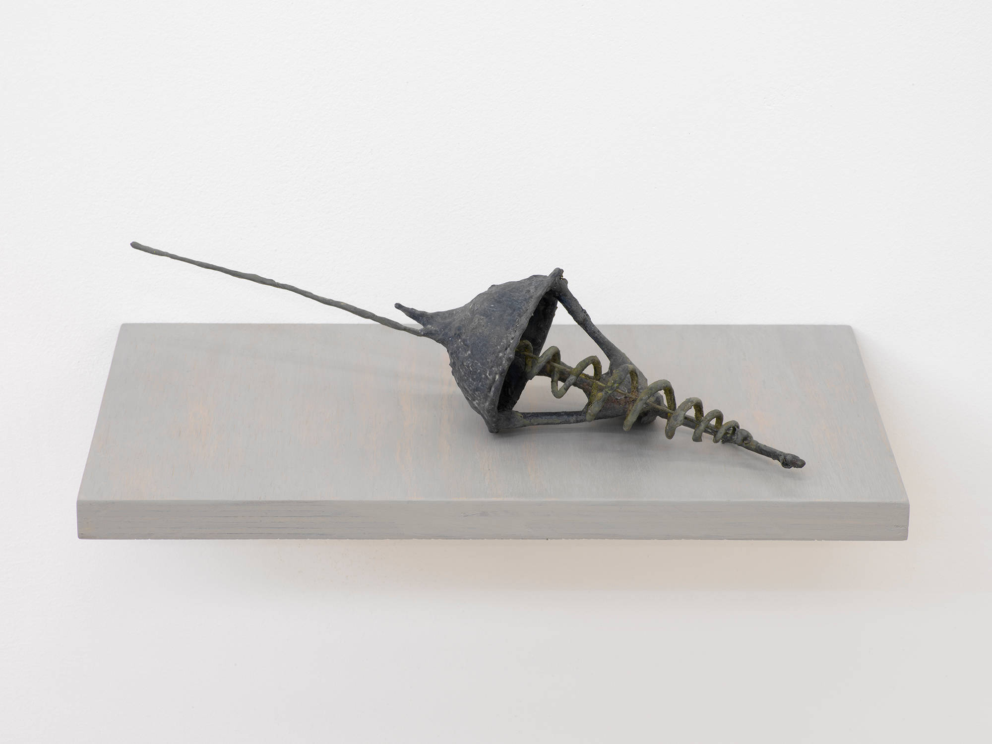 A small handmade abstract sculpture resembling a kind of artifactual tool or some combination of archaic materials. The object is resting on a gray shelf attached to a white wall.
