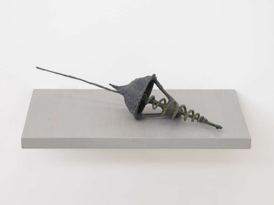 A small handmade abstract sculpture resembling a kind of artifactual tool or some combination of archaic materials. The object is resting on a gray shelf attached to a white wall.

